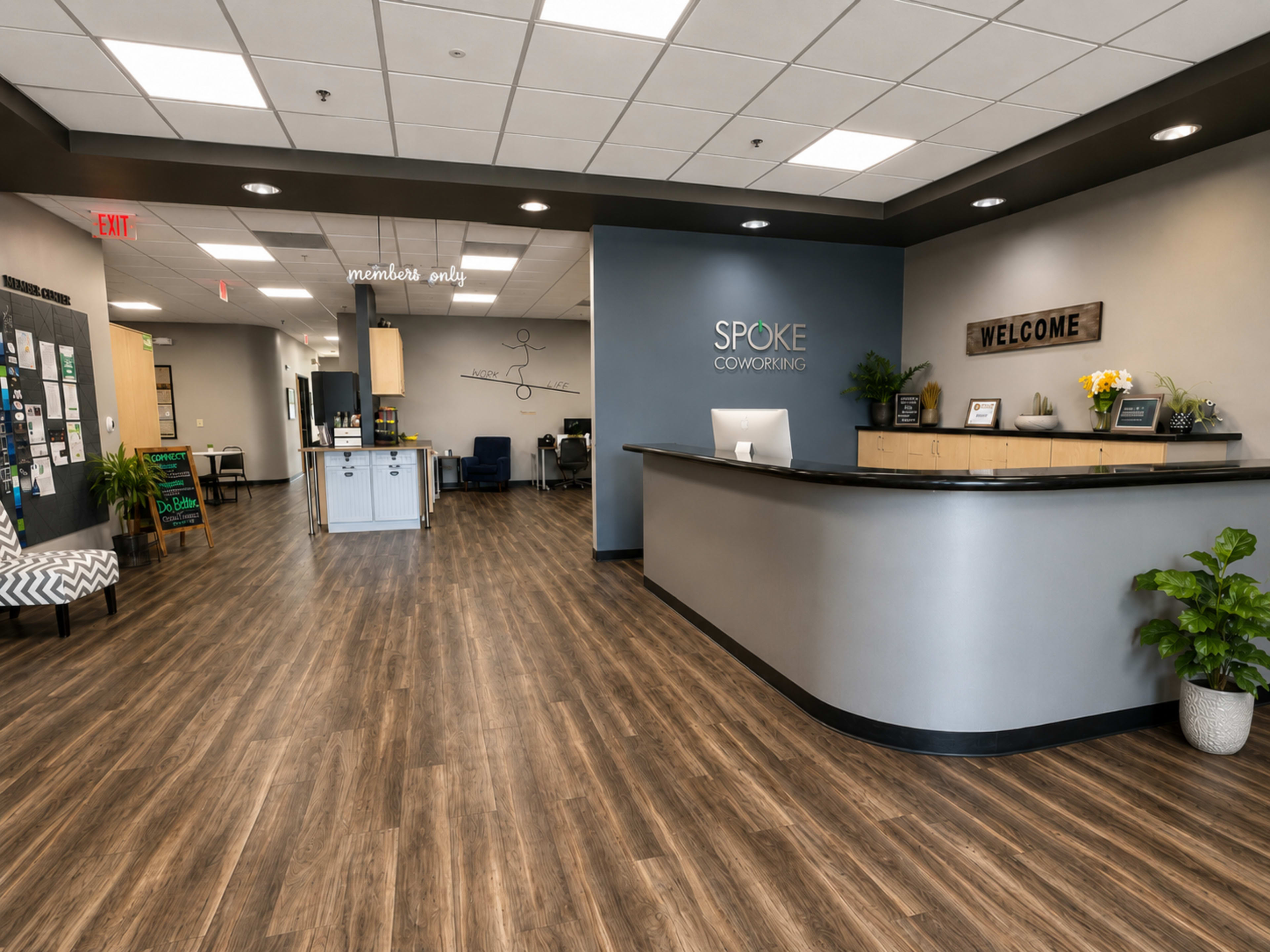 The image shows a modern coworking space reception area with a front desk, a welcome sign, and a seating area, featuring wooden flooring and neutral-colored walls.