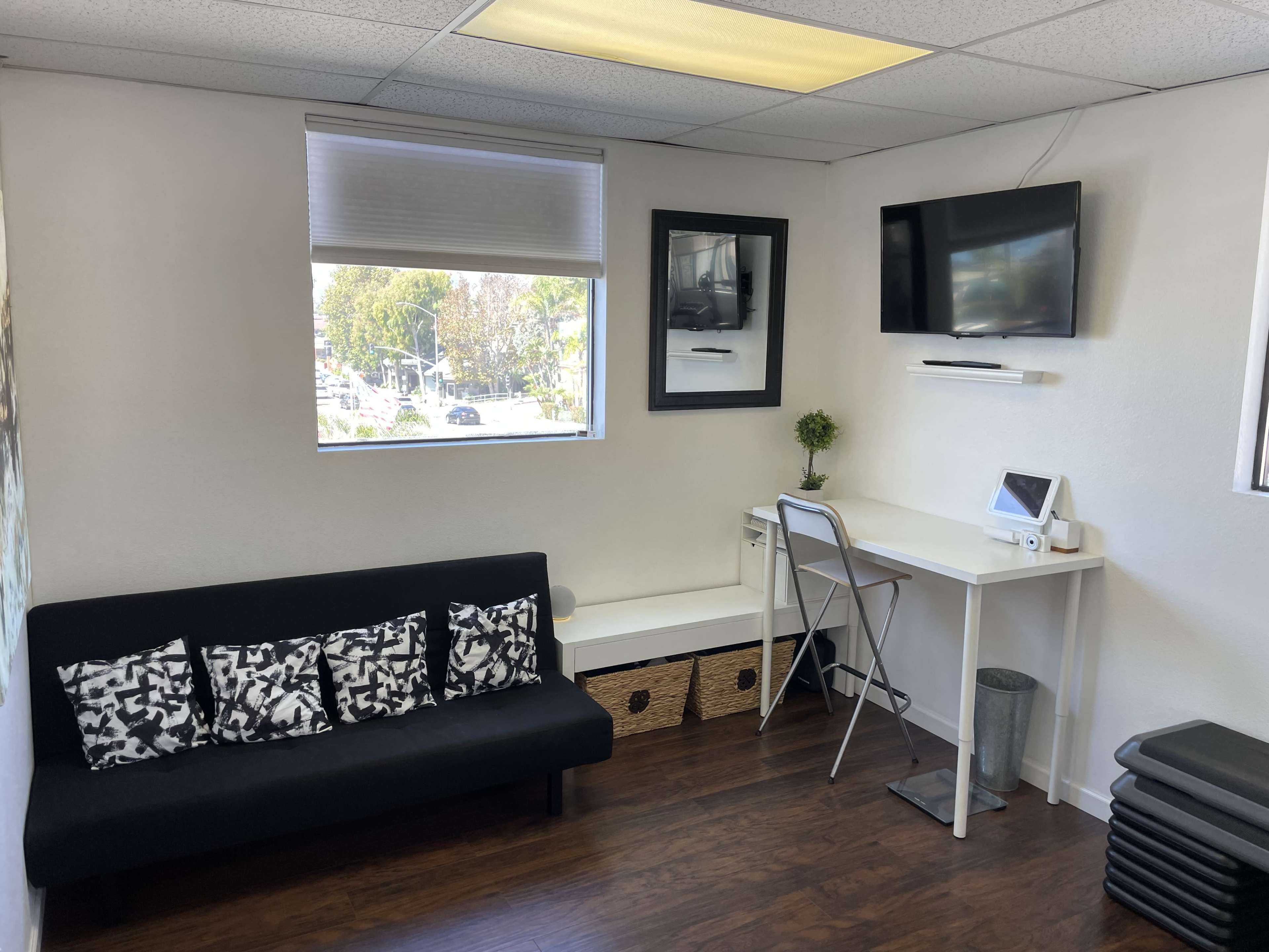 The image shows a small, modern office space with a black couch, a desk with a monitor, and a window overlooking a street.