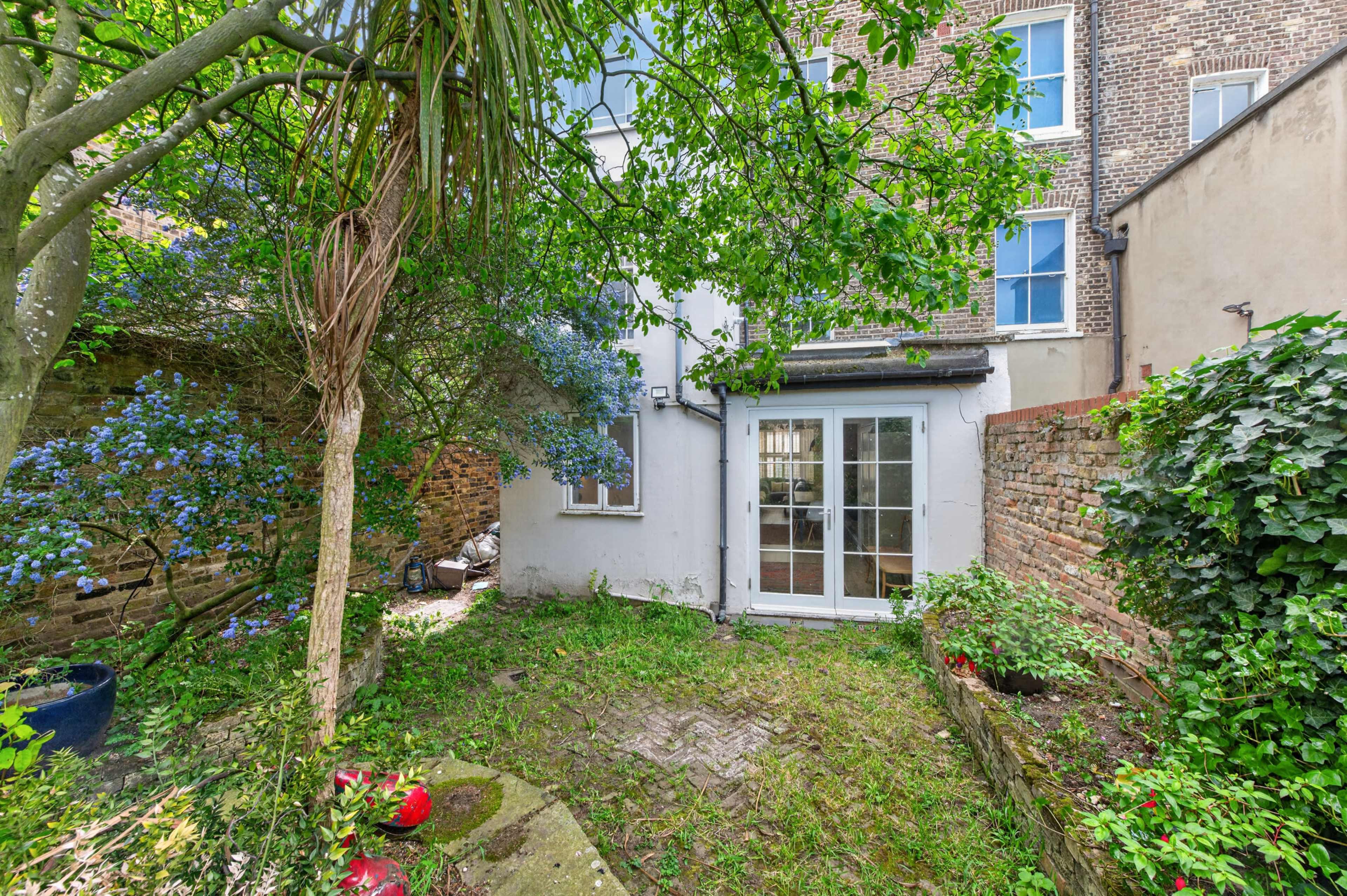 The image shows a small, overgrown garden with a brick wall, plants, and a set of French doors leading into a building.