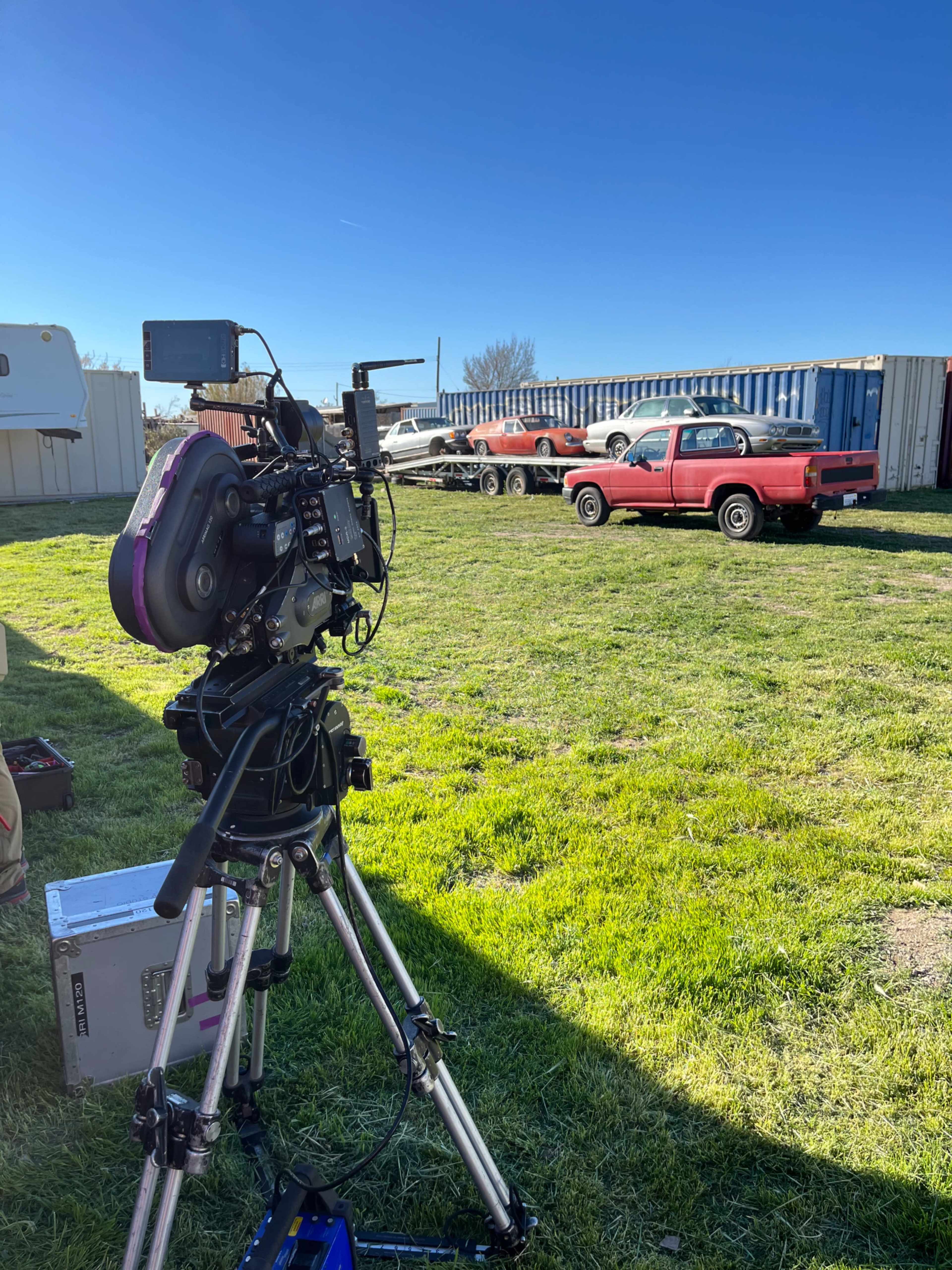 A camera setup on a tripod captures a scene of several parked cars and a cargo trailer in a grassy area under a clear blue sky.