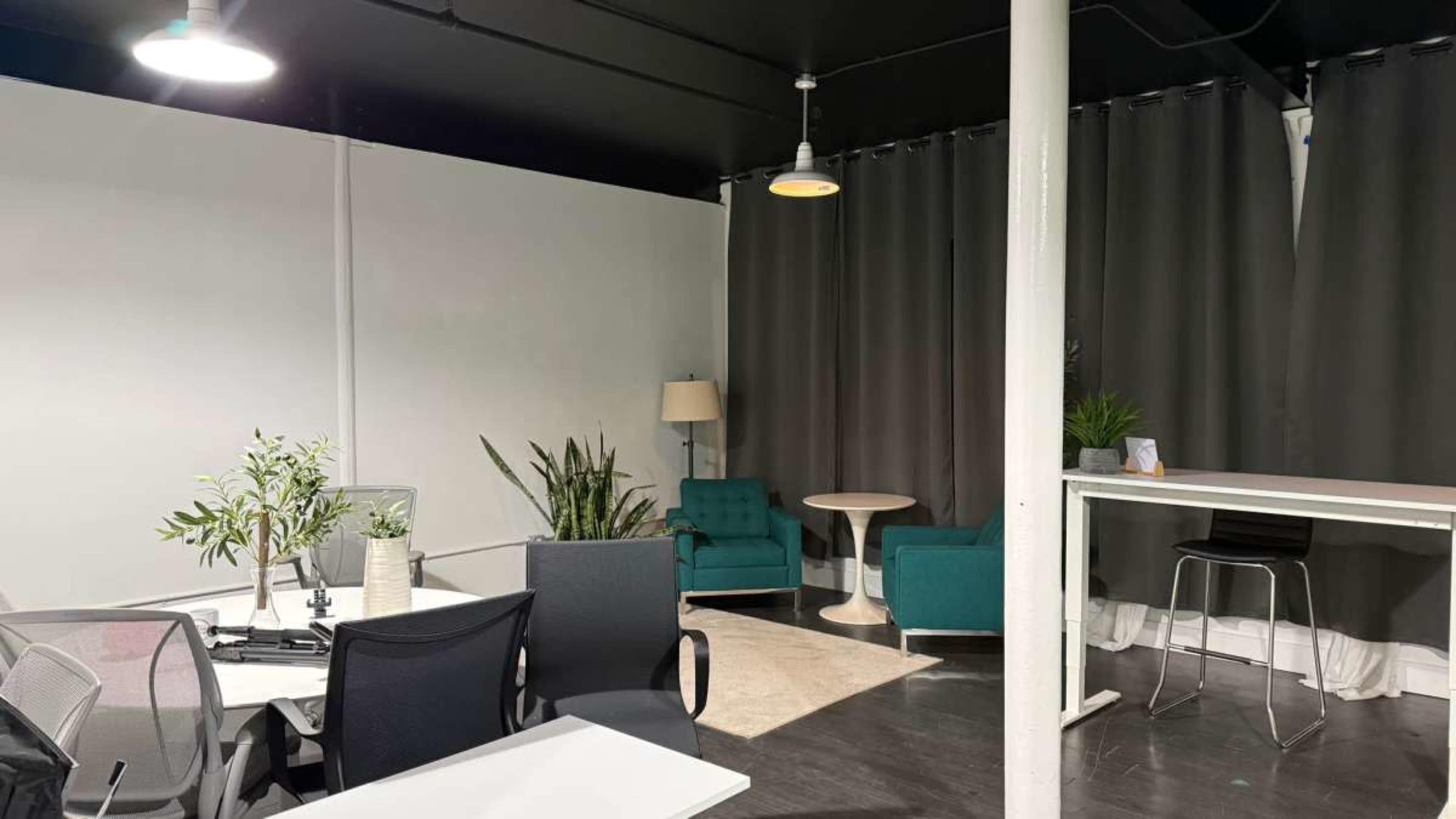 The image shows a modern office space featuring a seating area with two green chairs, a small table, and potted plants, alongside a workspace with multiple office chairs and desks.