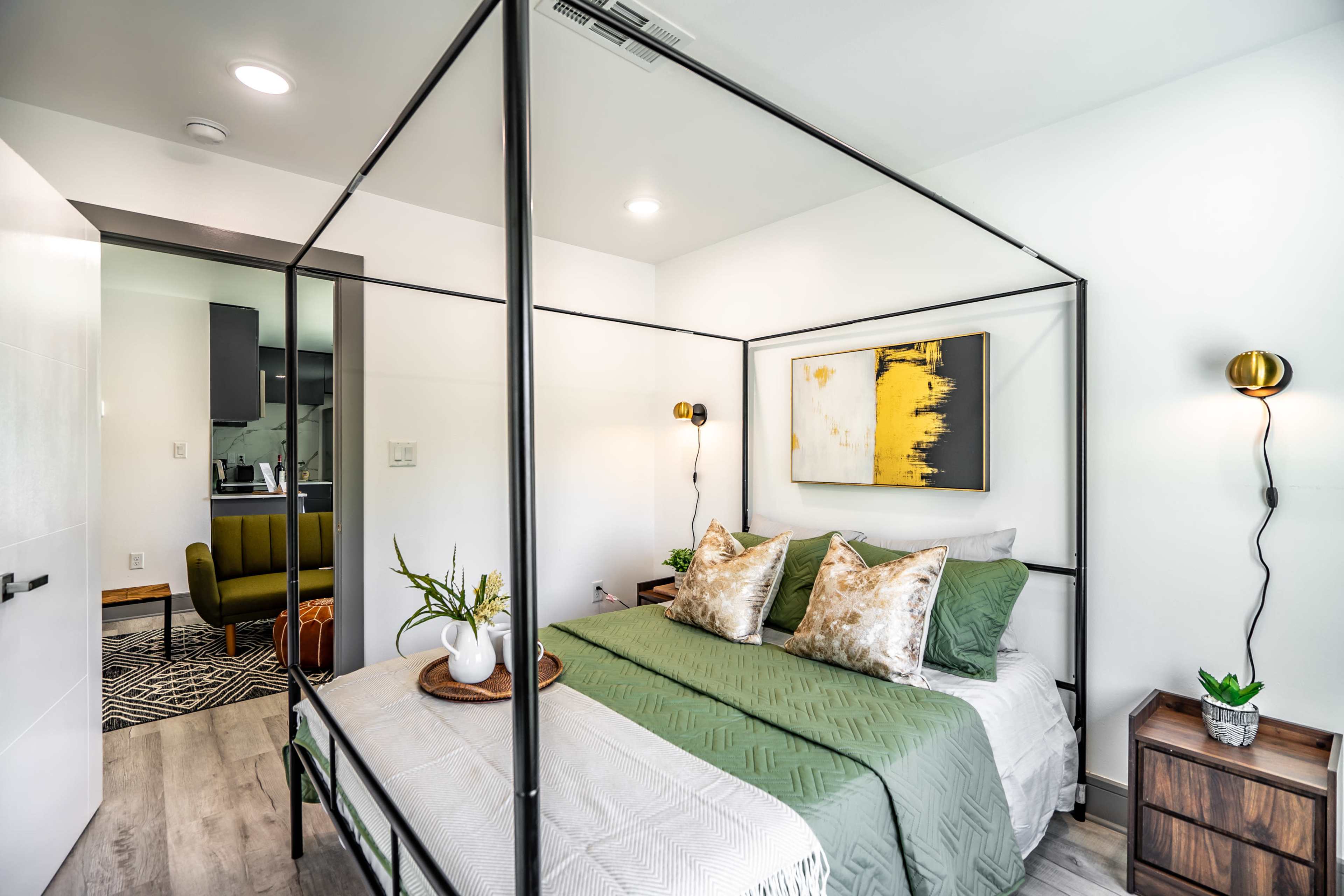 A modern bedroom features a four-poster bed with green bedding and decorative pillows, accompanied by bedside tables and a visible living area in the background.