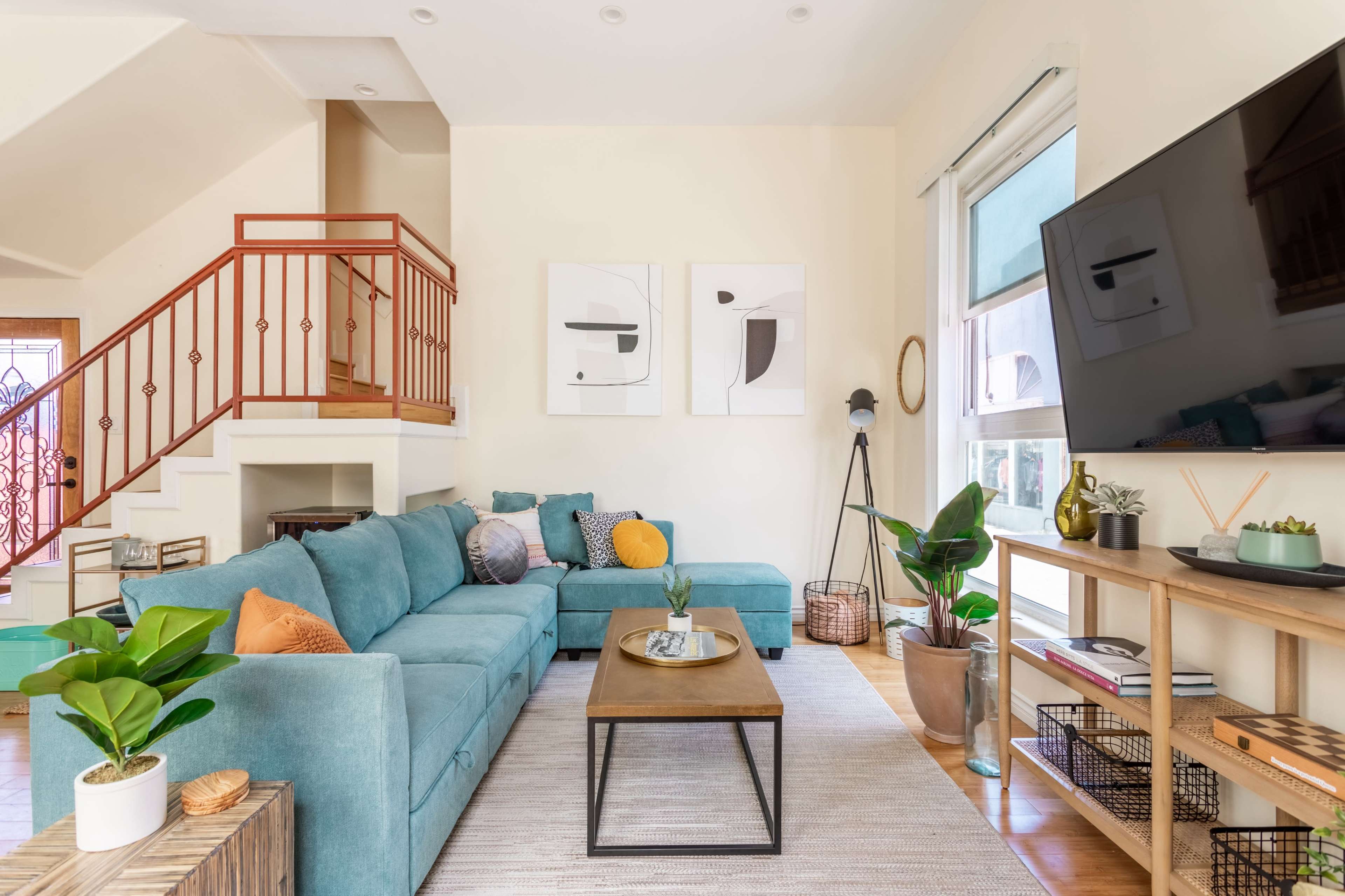 The scene features a modern living room with a teal sectional sofa, a wooden coffee table, and large windows, all complemented by minimalist artwork on the walls.