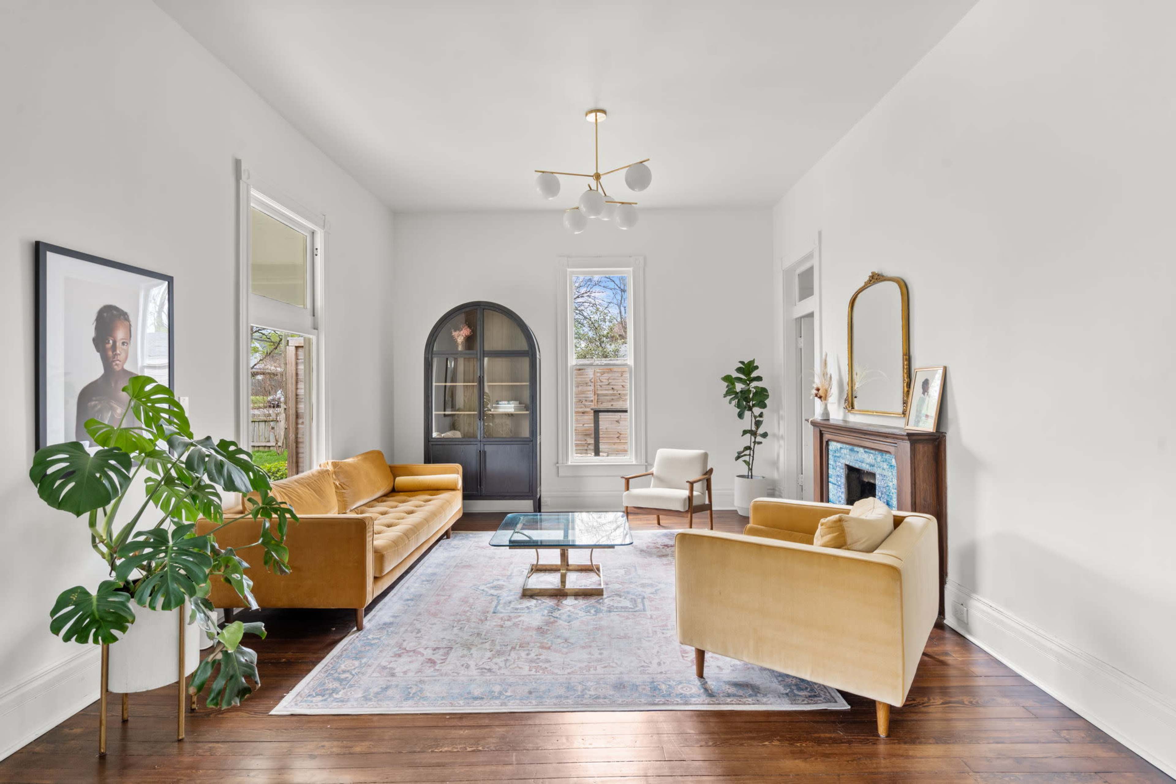 The image shows a spacious living room with a light-colored sofa, a glass coffee table, and a vintage fireplace, complemented by large windows and hardwood flooring.