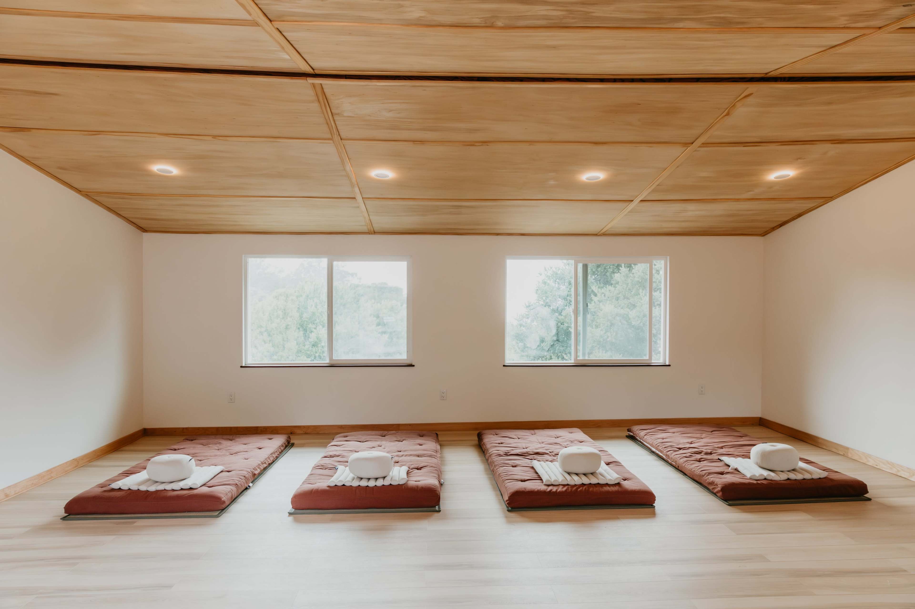 The image shows a serene room with four futon mattresses laid out on a wooden floor, accompanied by round cushions, and large windows overlooking greenery.