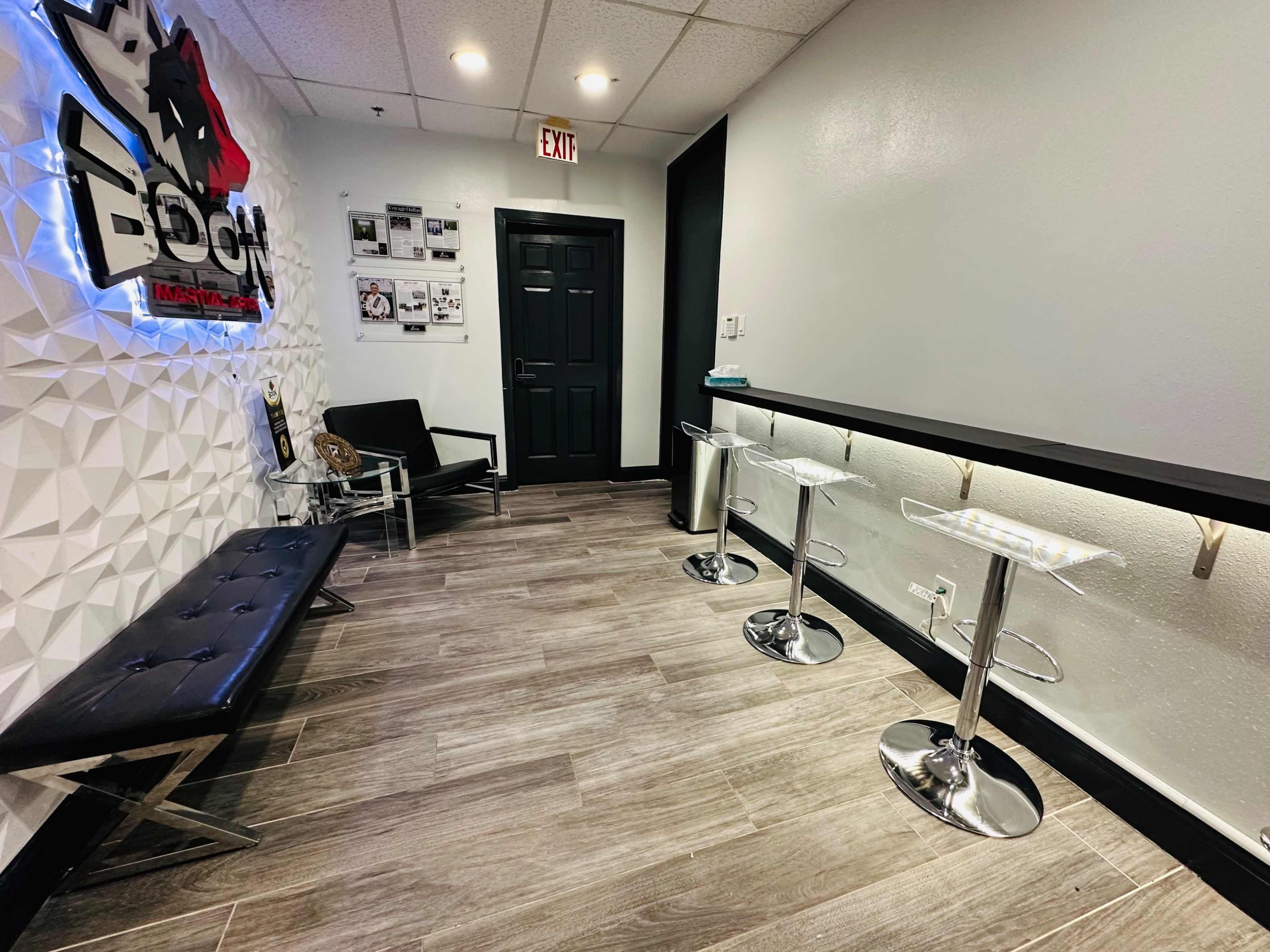 A modern waiting area with a black bench, two bar-style tables, and a wall adorned with various documents and a lighted sign.