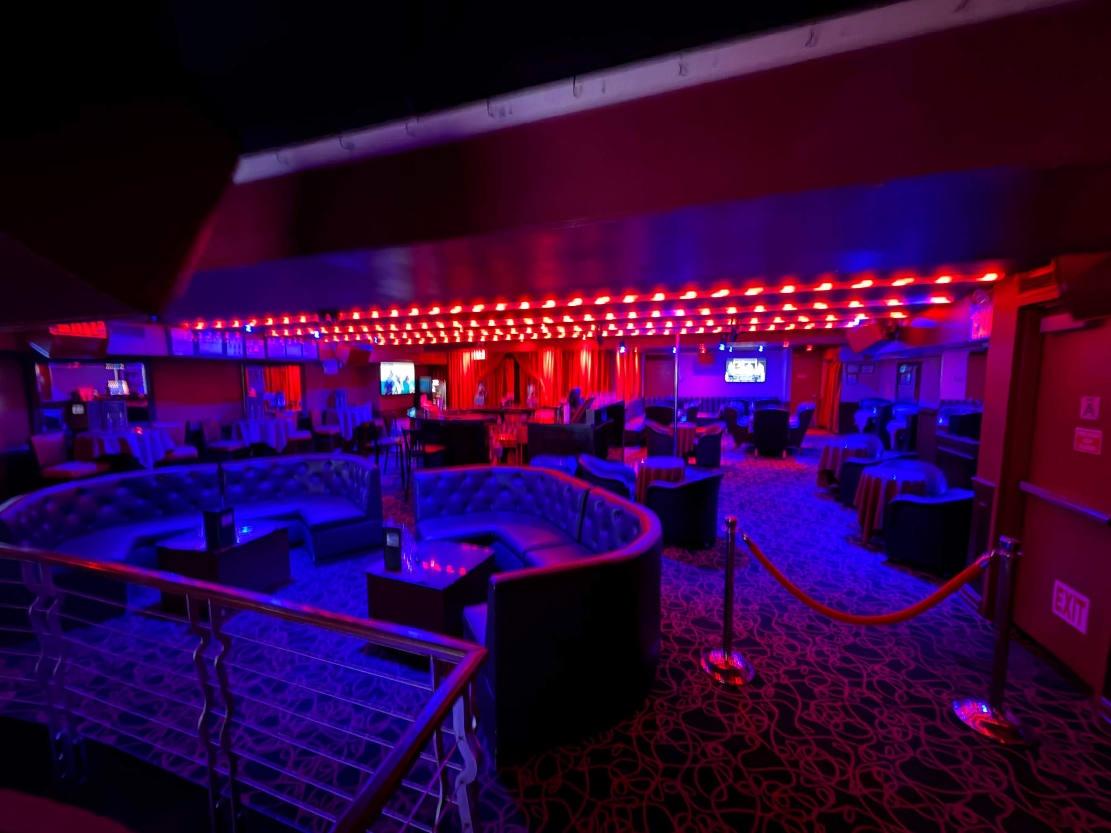The image shows a dimly lit lounge with blue and red lighting, featuring circular seating arrangements and small tables throughout the space.