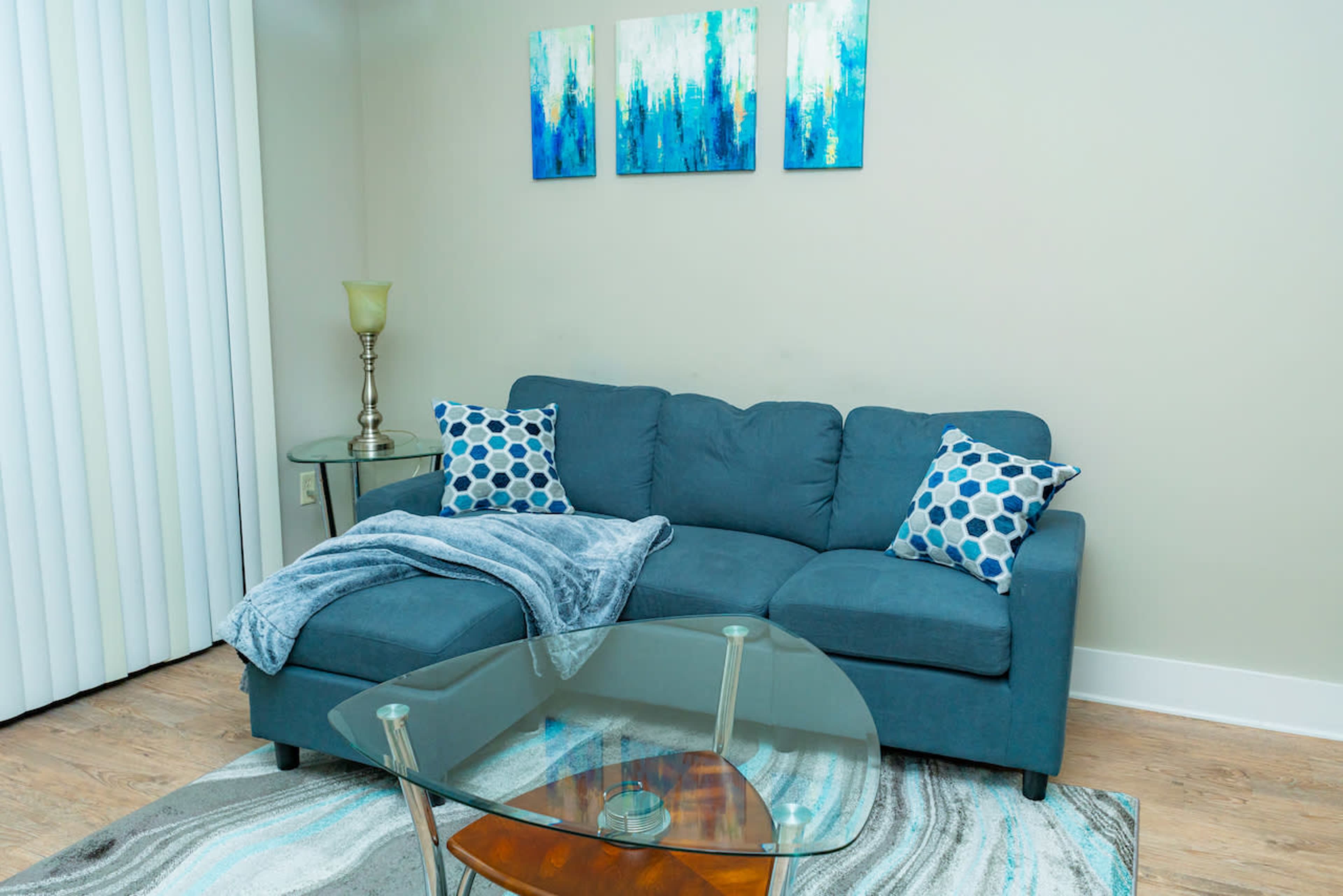 A blue couch with patterned pillows is positioned next to a glass coffee table on a patterned rug beneath three decorative blue and green paintings.