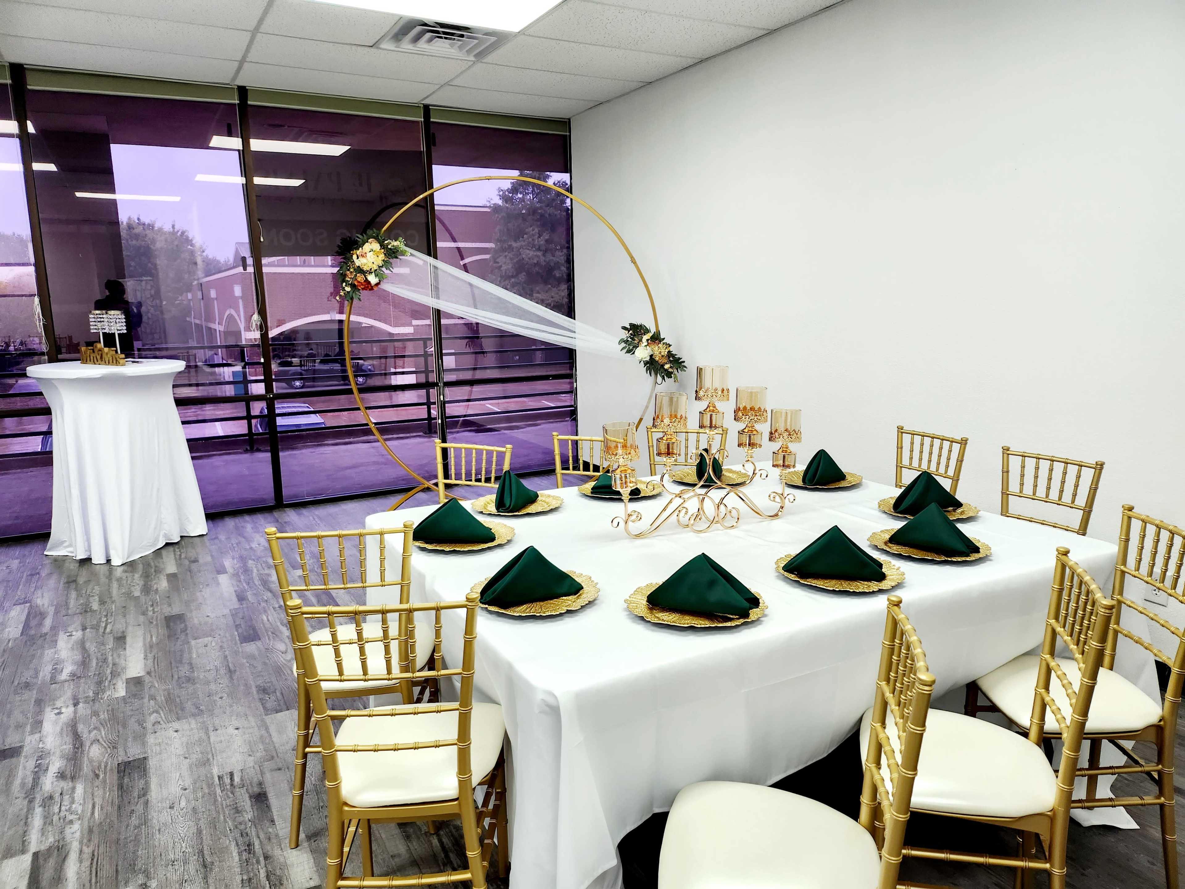 A elegantly arranged dining setup features a table with a white tablecloth, gold chairs, green napkins, and decorative centerpieces in a well-lit room with large windows.