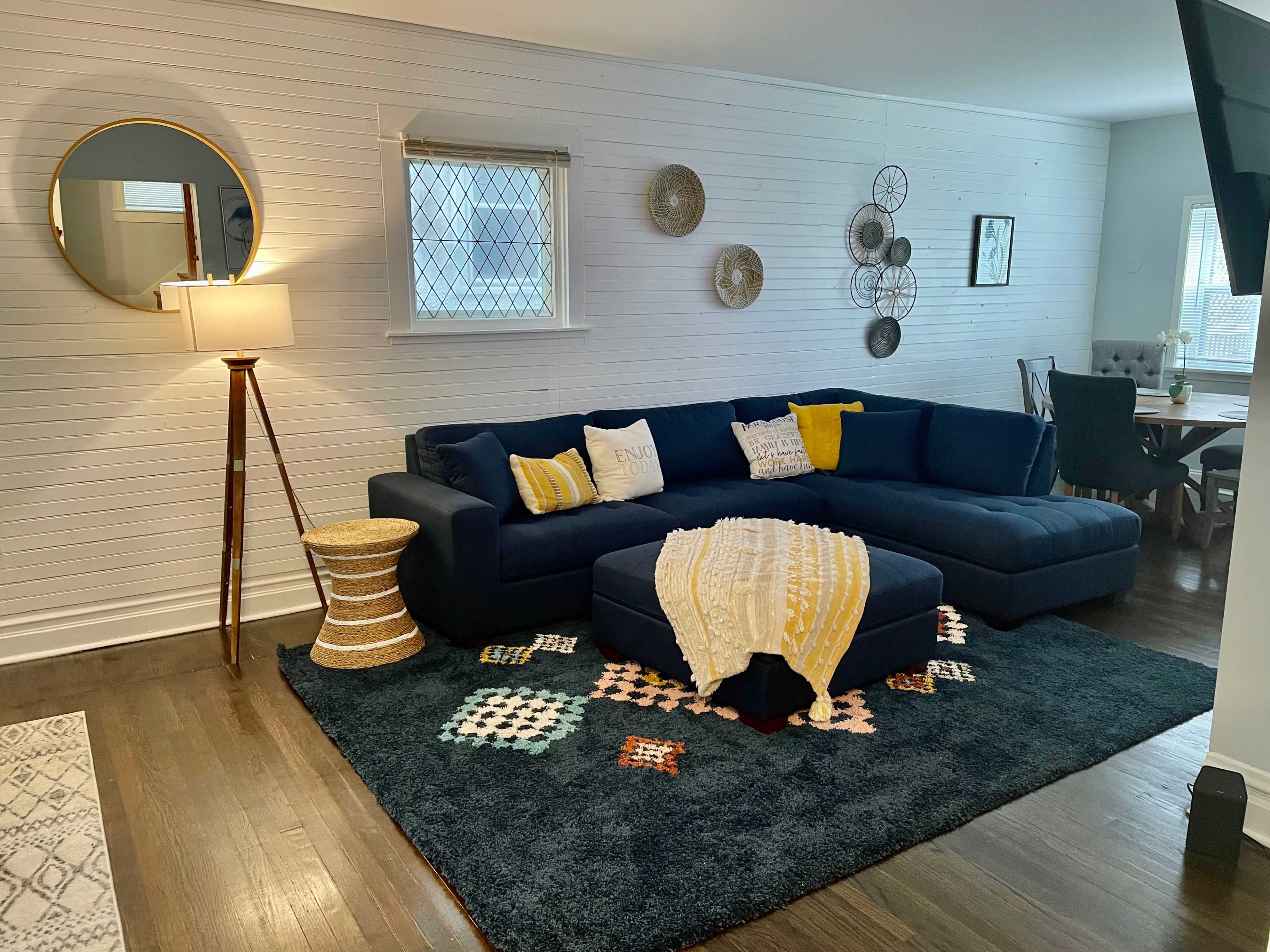 A cozy living room featuring a dark sectional sofa, a round mirror, a lamp on a wooden floor, and decorative wall hangings, all arranged around a textured rug.