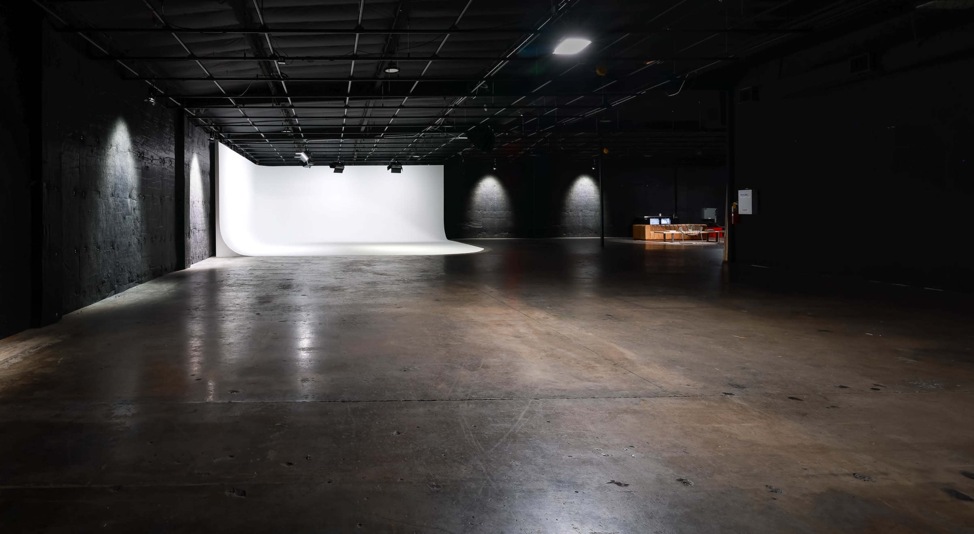 The image shows a spacious studio with a white backdrop at one end and a reception area with a desk at the opposite end, illuminated by overhead lights.