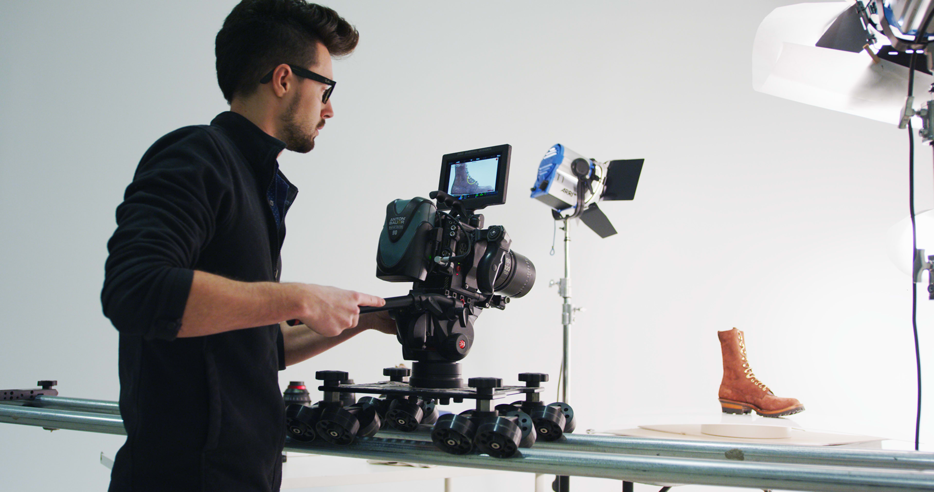 A camera operator adjusts a professional camera on a slider while capturing a close-up of a brown boot in a studio setting.