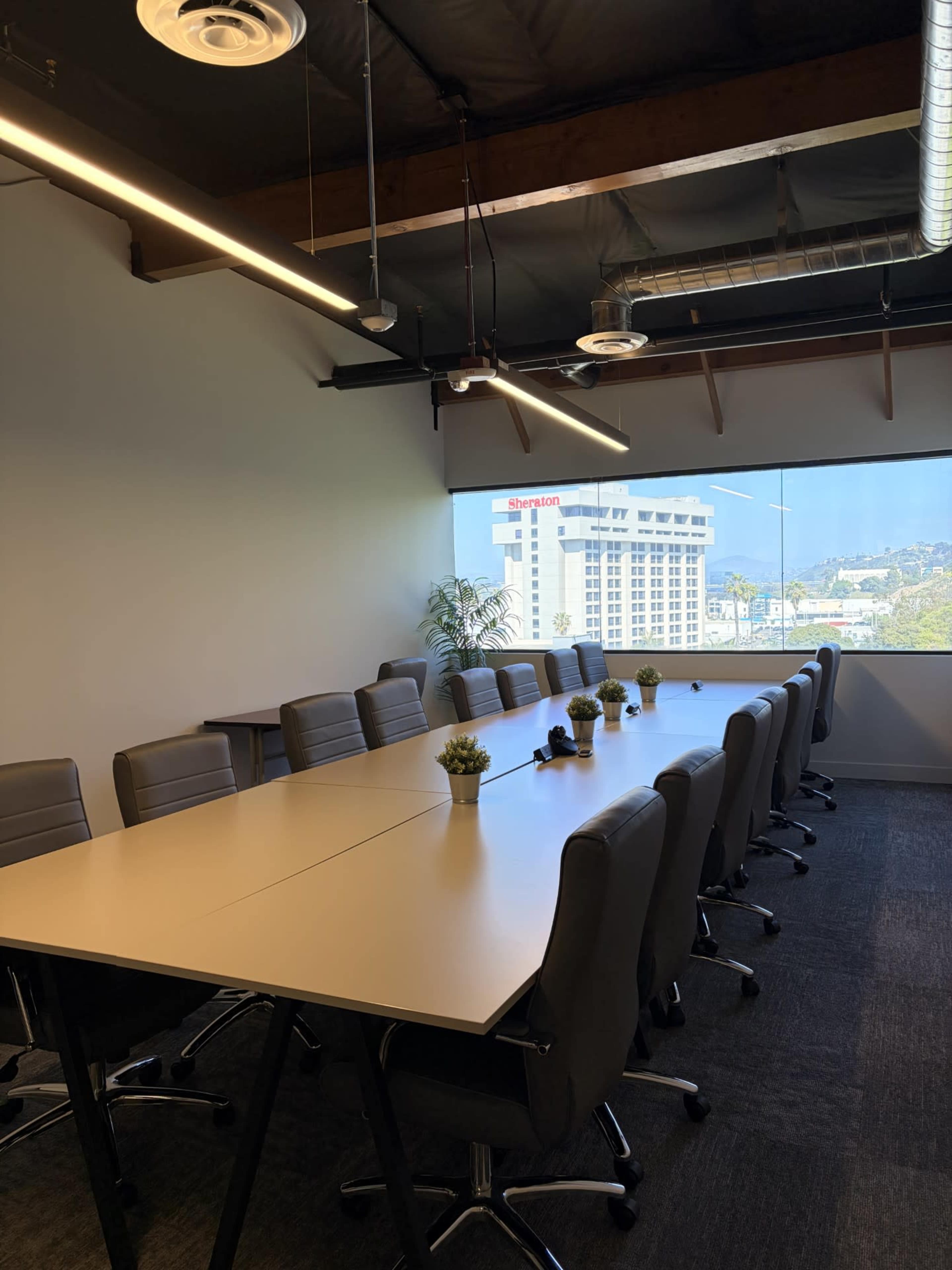 A large conference room features a long table surrounded by ergonomic chairs, with a view of a cityscape through a window.