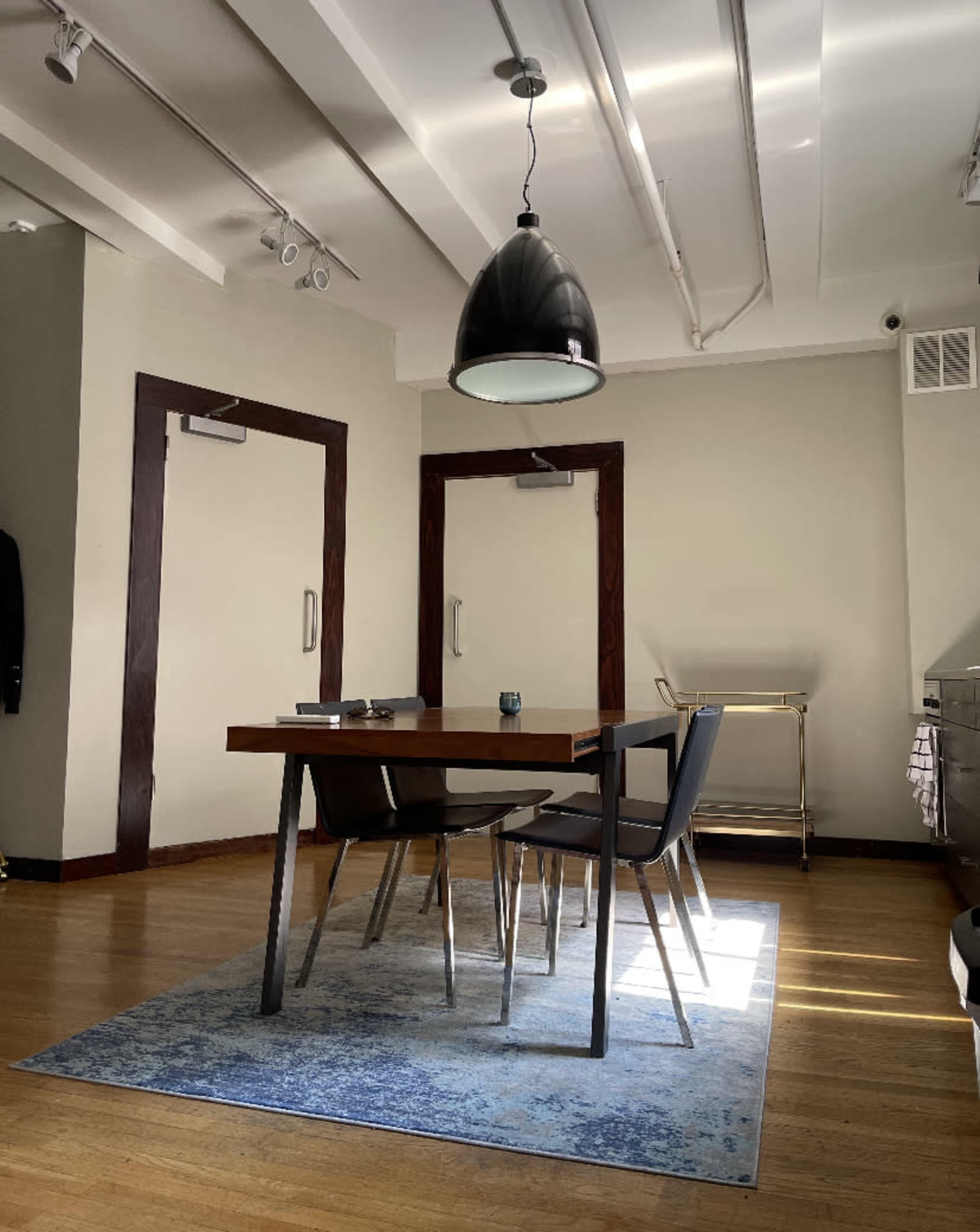 A modern dining area features a wooden table with four black chairs on a blue rug, illuminated by a pendant light.