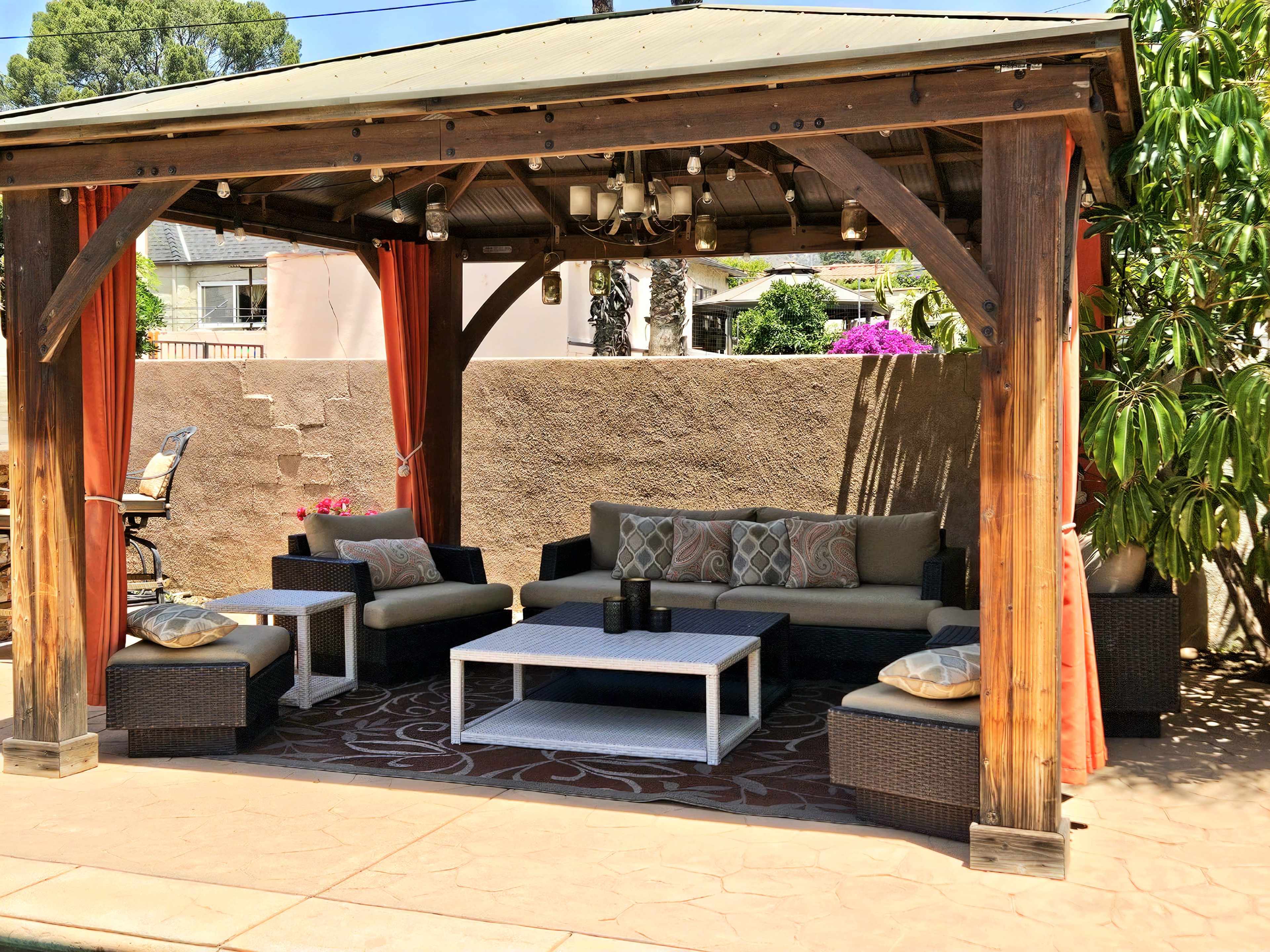 The image shows a shaded outdoor seating area with a wooden pergola, featuring a grey sofa set, a white coffee table, and potted plants.