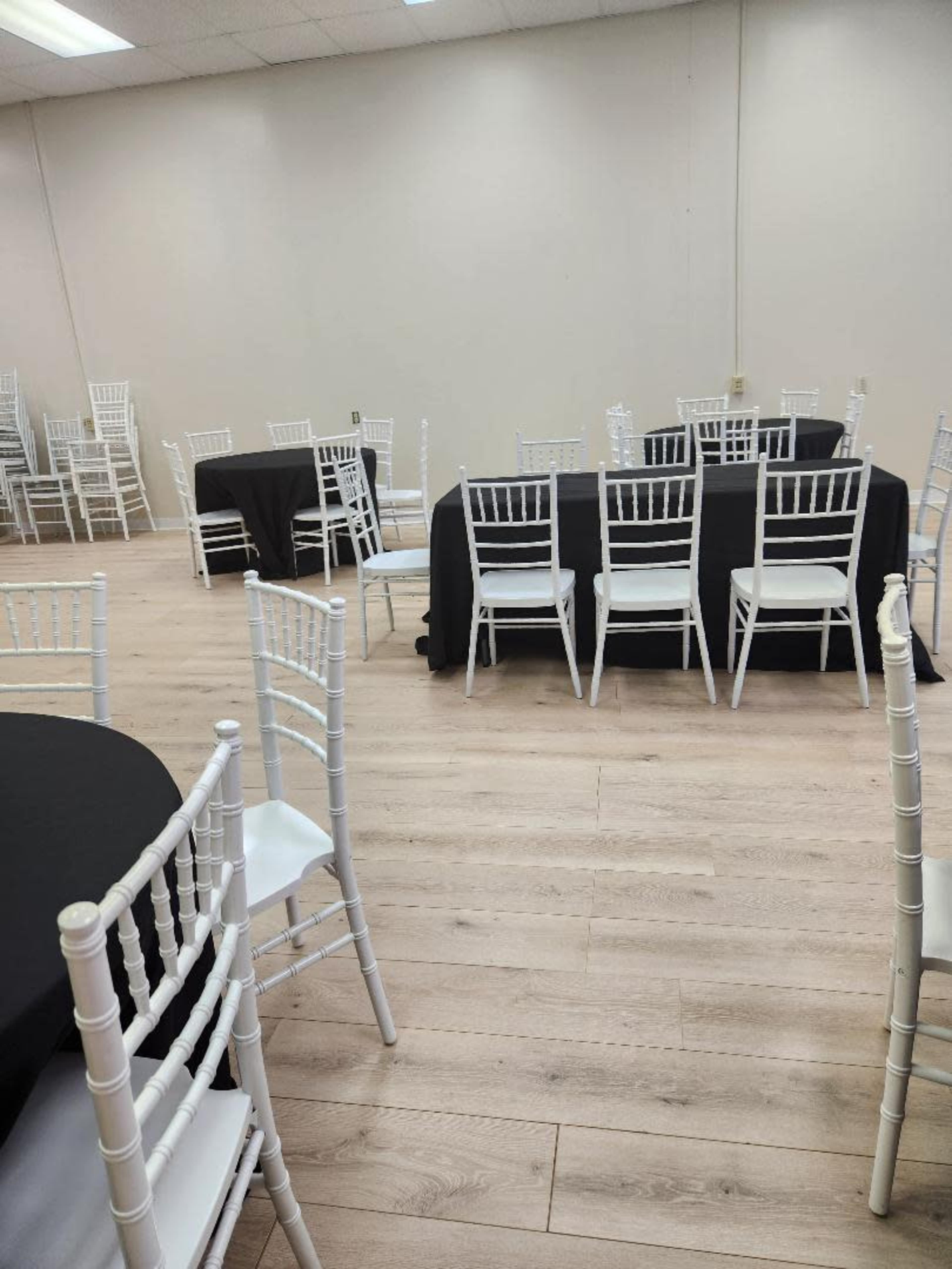 A spacious event room with white chiavari chairs arranged around black and white tables on a wooden floor.