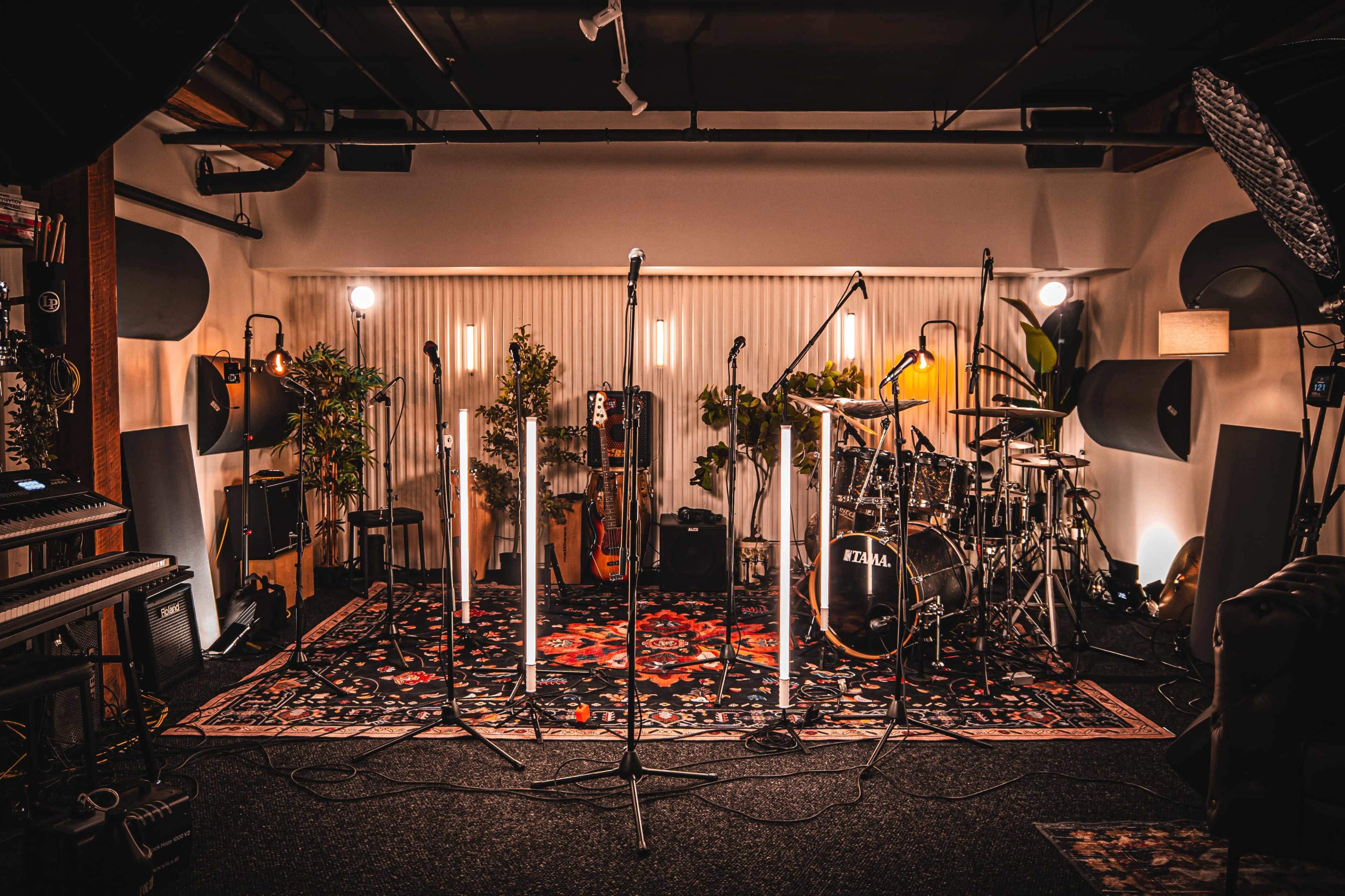 The image shows a music studio set up with microphones arranged in a circle around a drum kit, surrounded by plants and lighting fixtures.