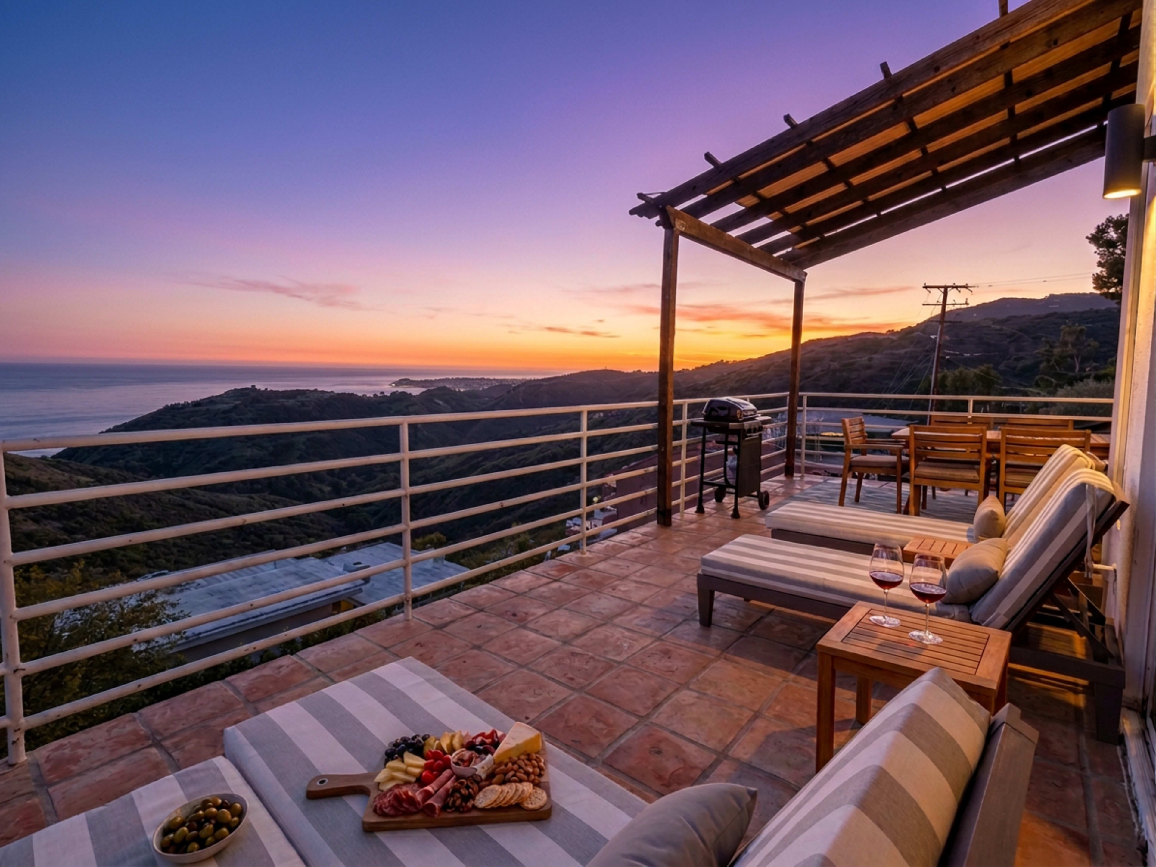 The image shows a terrace with lounge seating and a dining area, overlooking a coastal view at sunset.