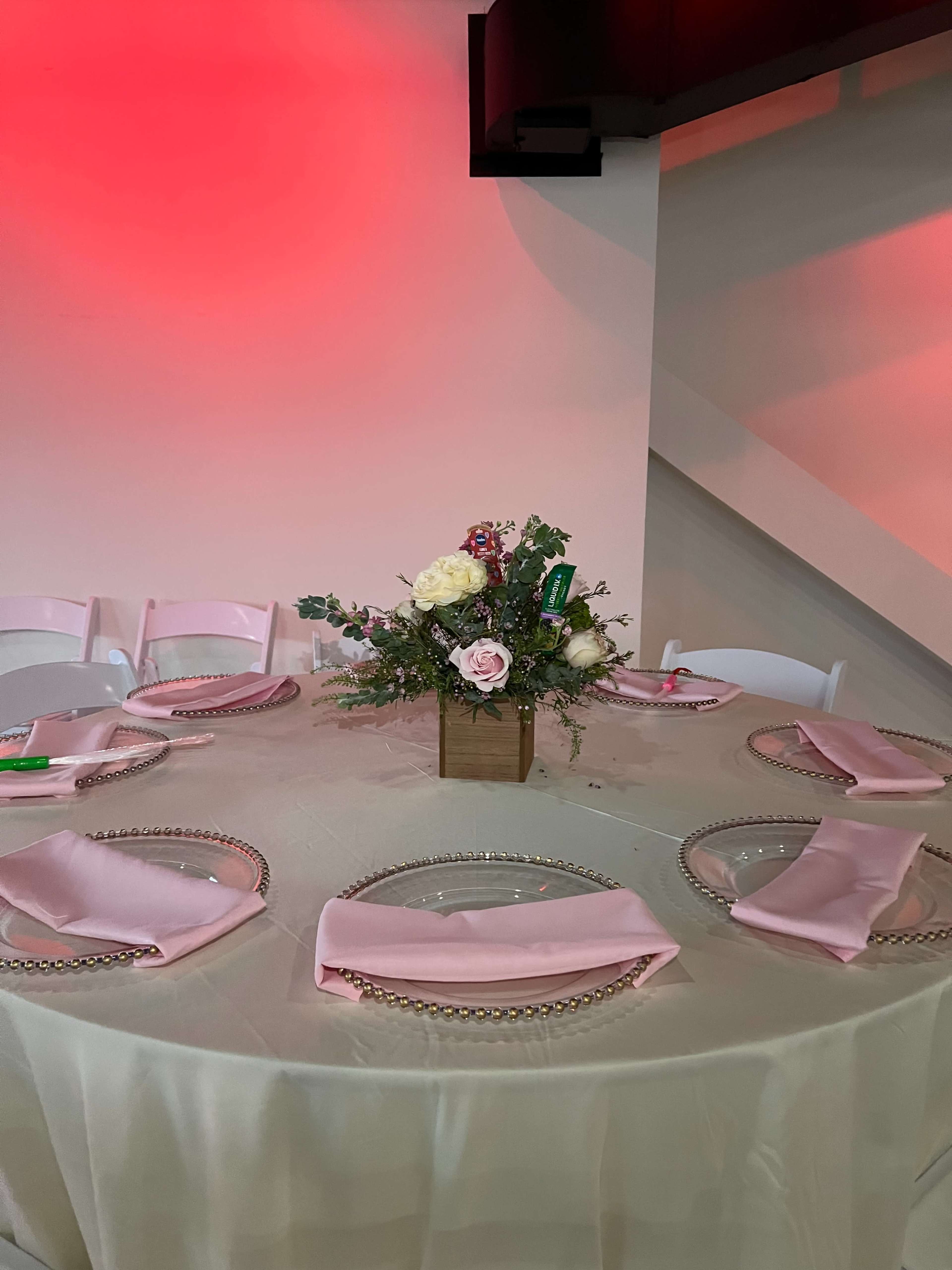 A round dining table is set with pink napkins, decorative plates, and a floral centerpiece, all under soft red lighting.