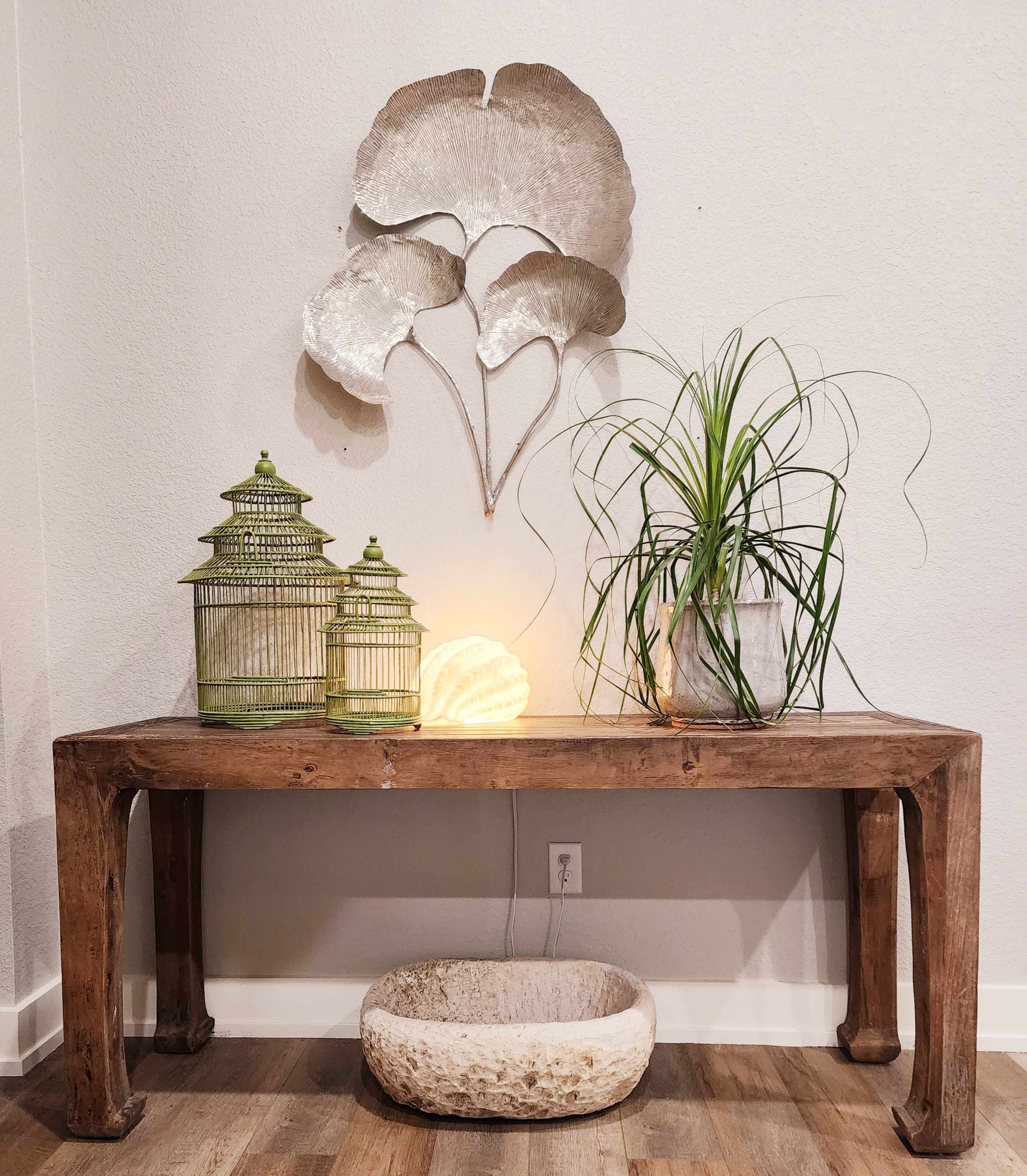 A wooden console table is adorned with decorative green birdcages, a potted plant, a glowing orb, and a wall-mounted metallic decor piece resembling leaves.