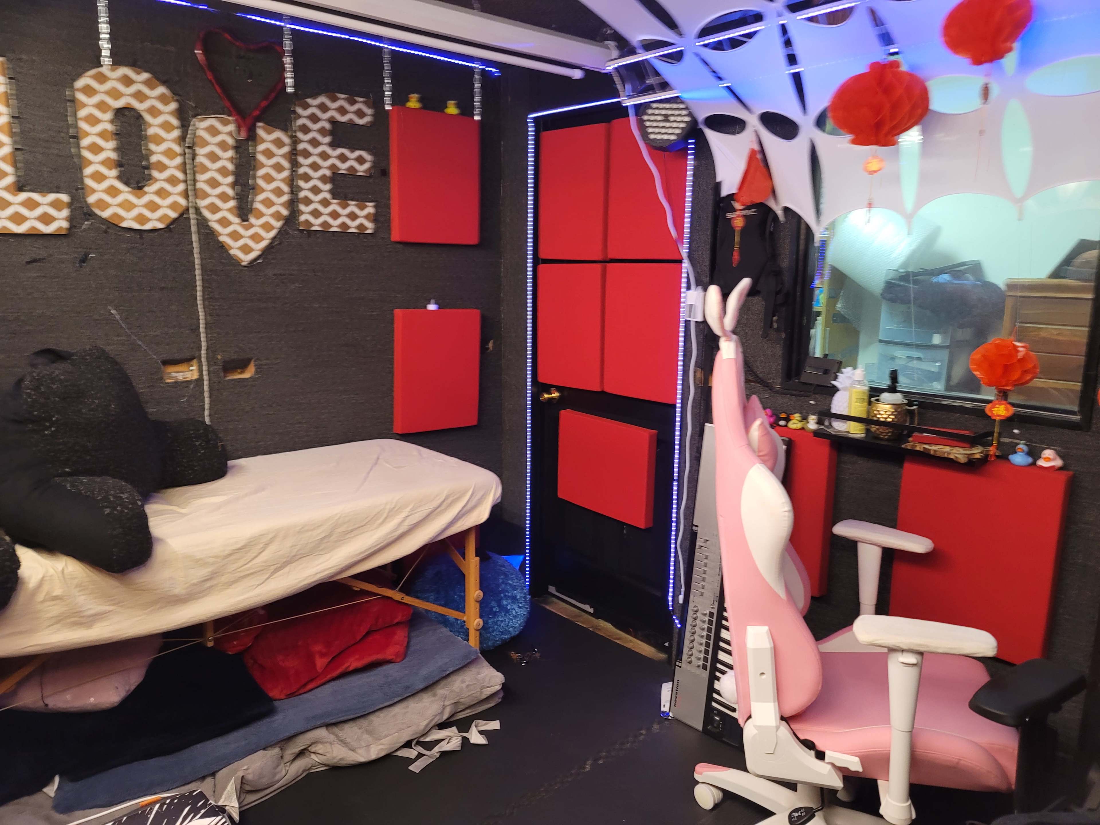 A small room with a bed, a pink gaming chair, and walls adorned with decorations and red panels.