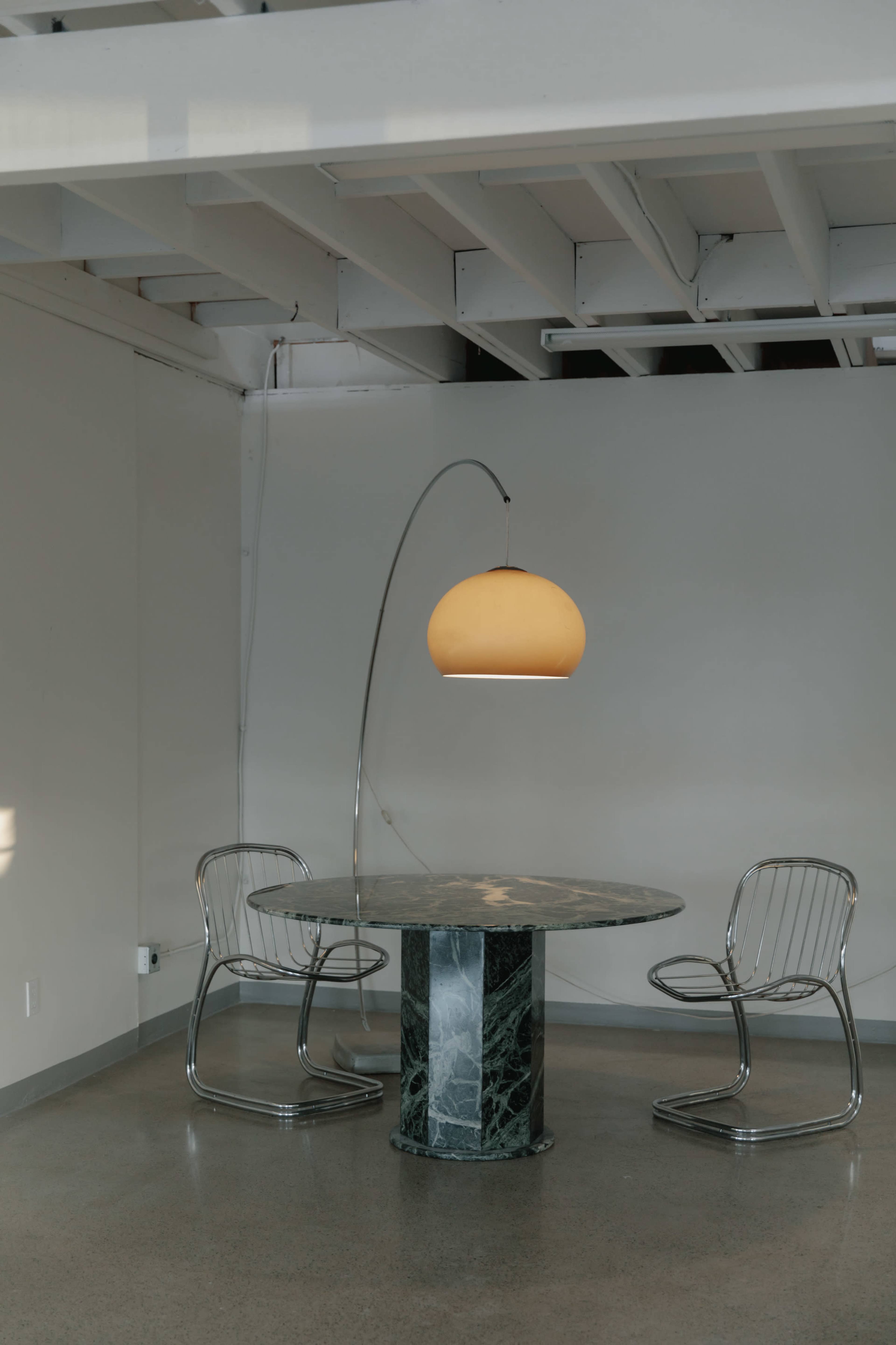 A round marble table with a curved metallic base is paired with two wireframe chairs under a large, arched pendant lamp in a minimalist room.