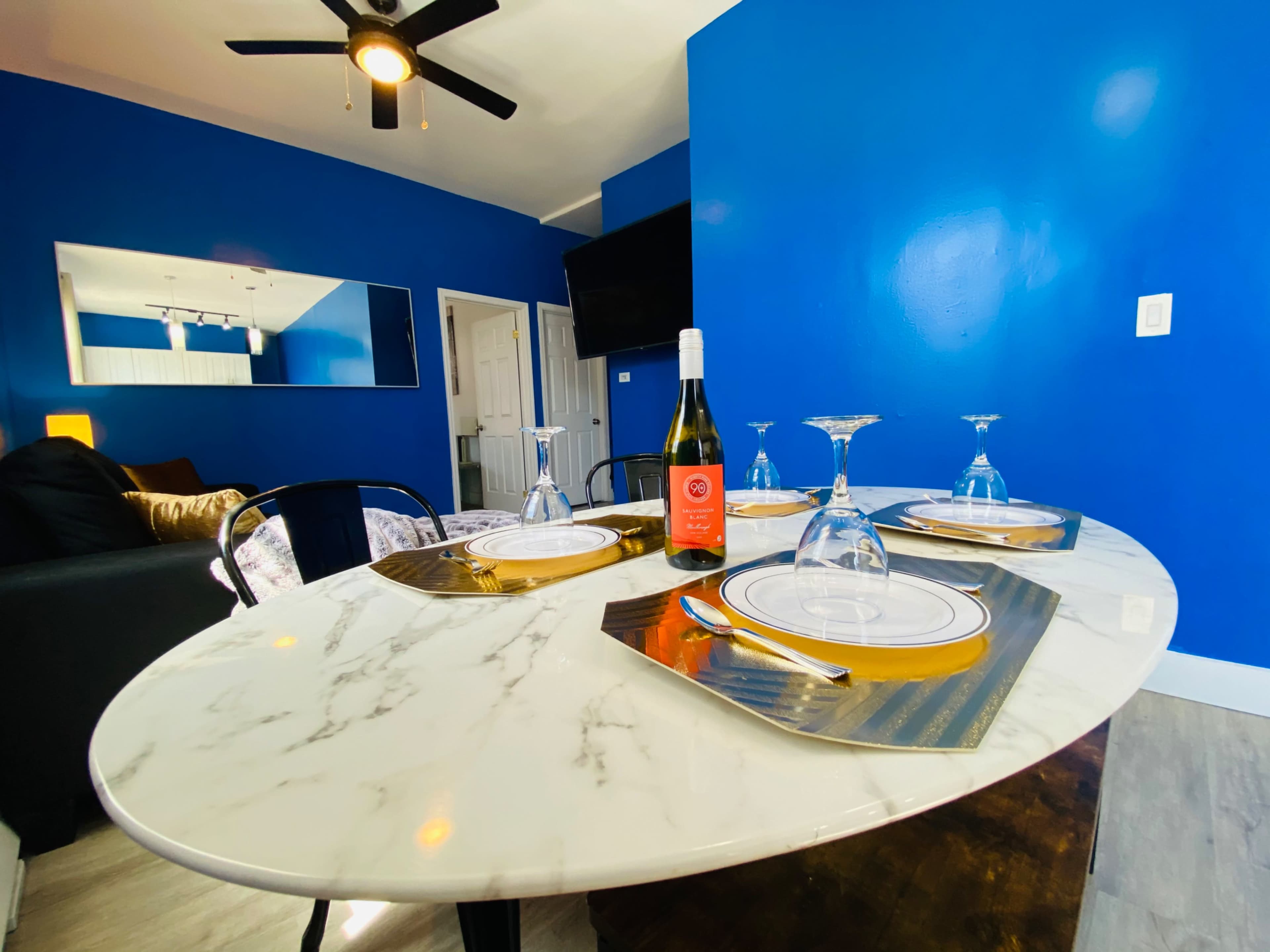 A circular marble dining table is set with wine glasses and plates, surrounded by a blue-walled room featuring a mirror and a television.