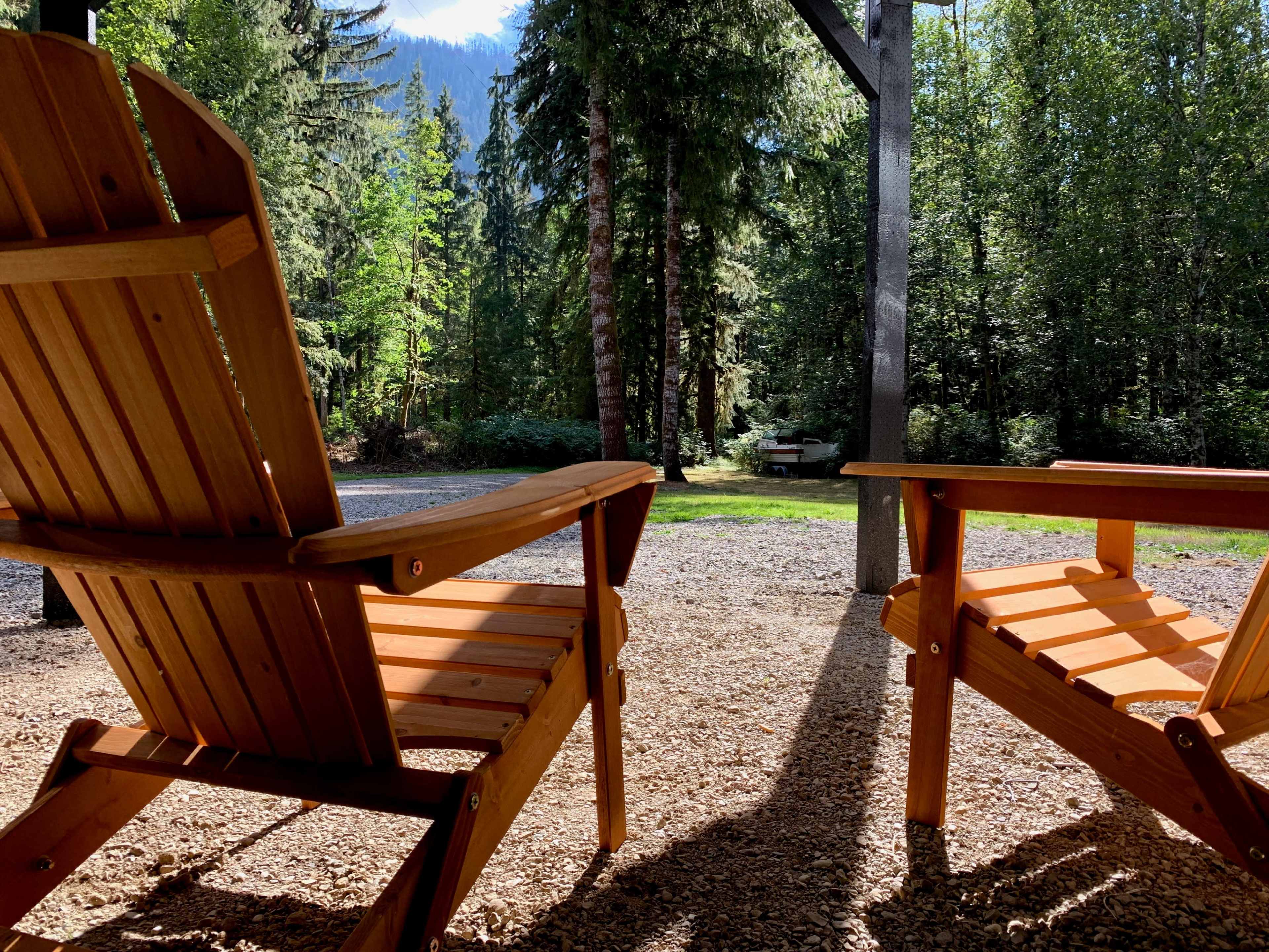 Two wooden chairs are positioned under a shaded porch, overlooking a gravel pathway surrounded by tall trees.