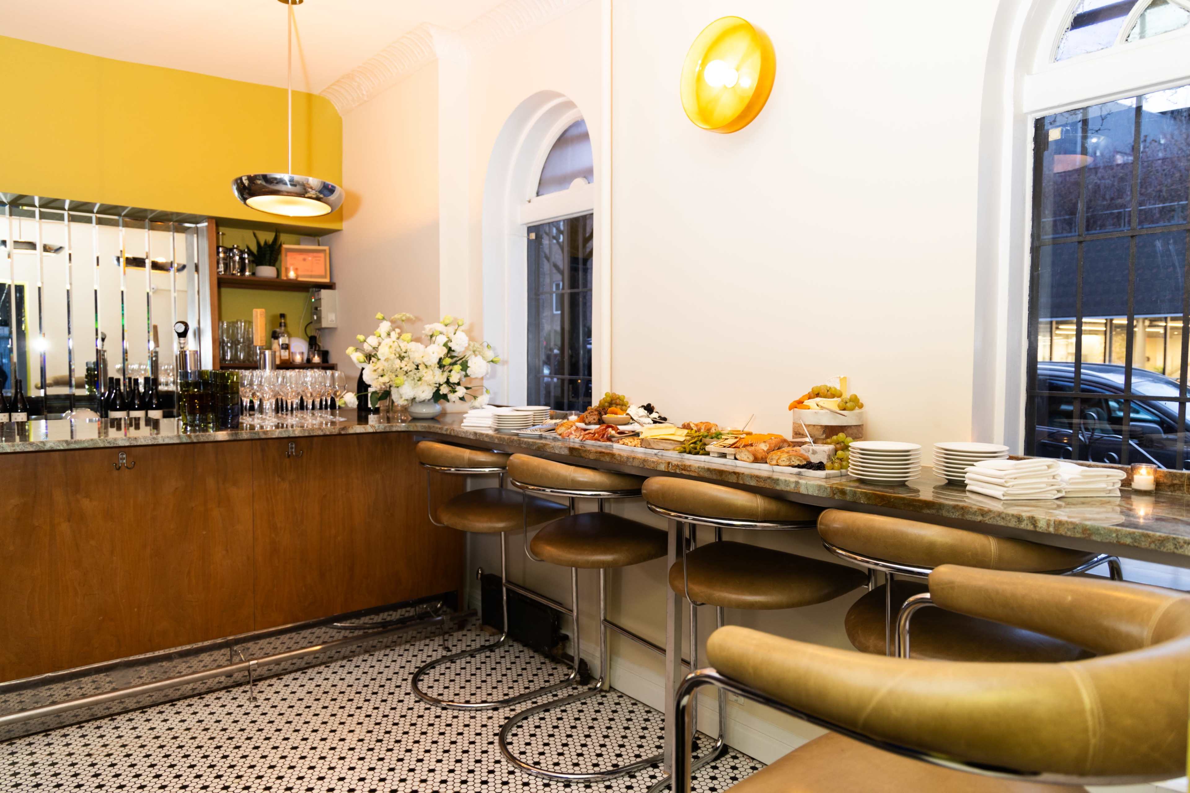 A stylish bar area features a long marble counter with comfortable stools, a display of food, and shelves stocked with drinks and floral arrangements.
