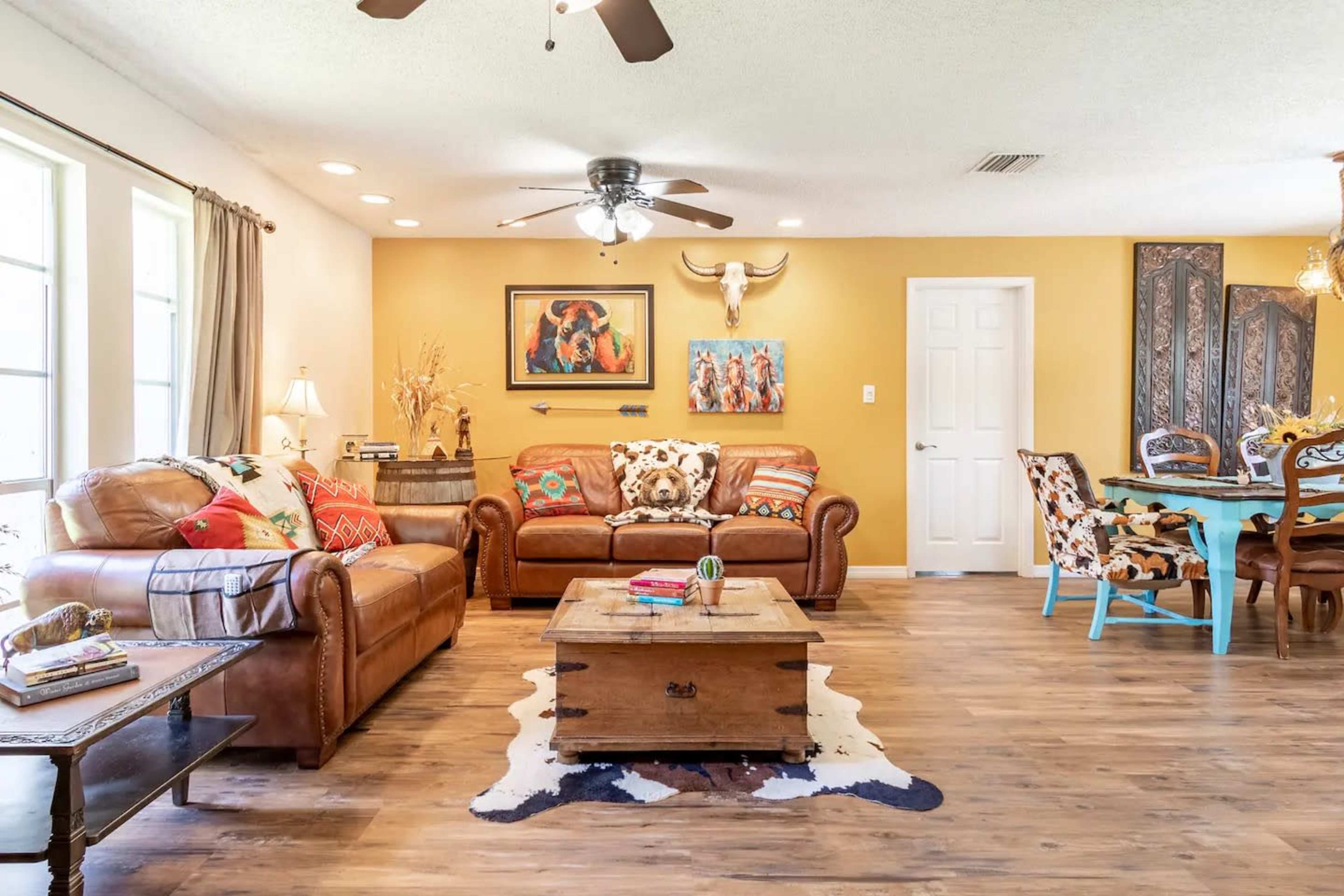 The living room features brown leather sofas, a wooden coffee table, and a dining area with a colorful table and chairs, all set against a warm yellow accent wall.
