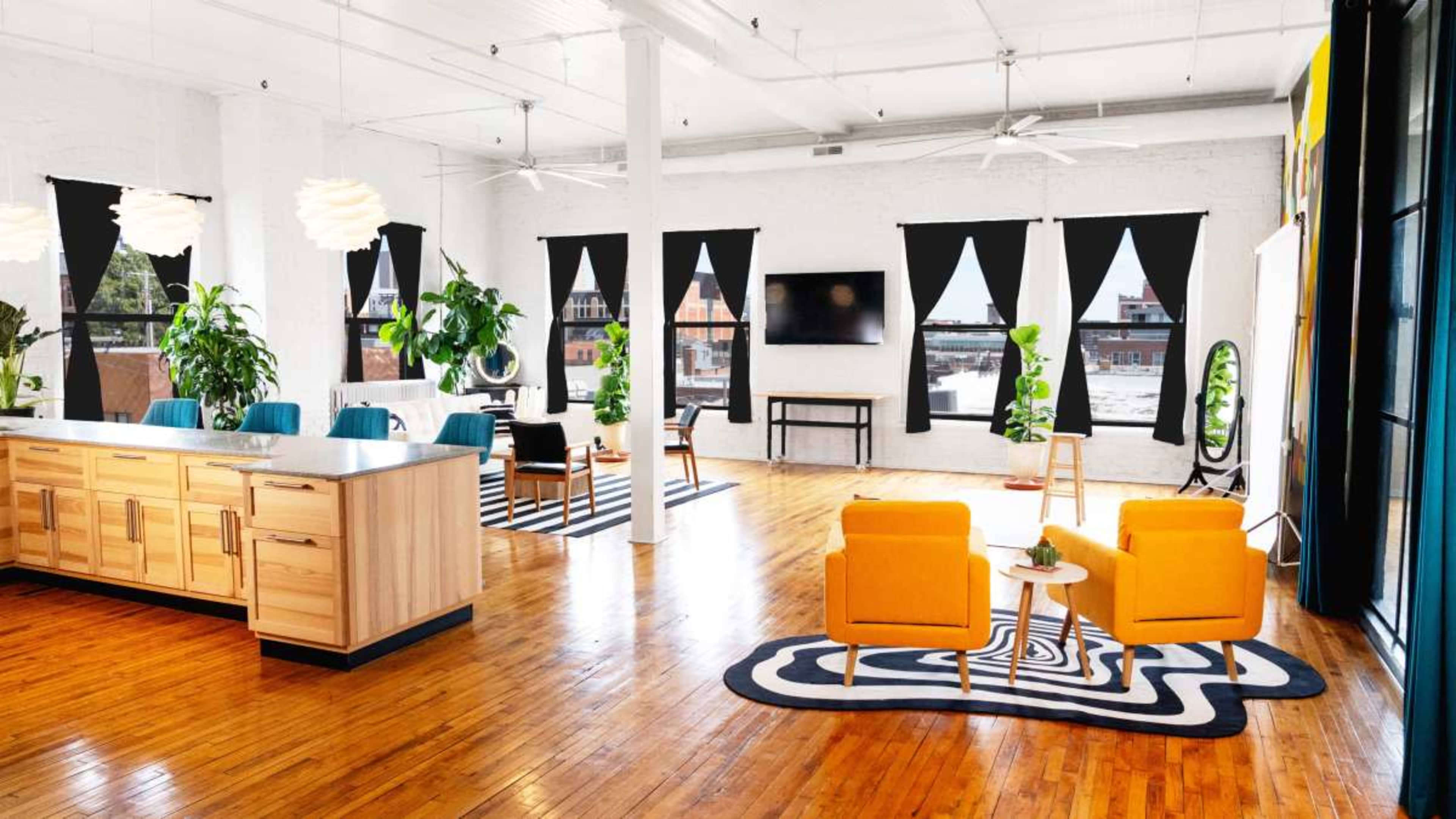 The image shows a spacious, modern indoor area with wooden flooring, large windows, and various plants, featuring a kitchen island and seating arrangements with orange chairs.
