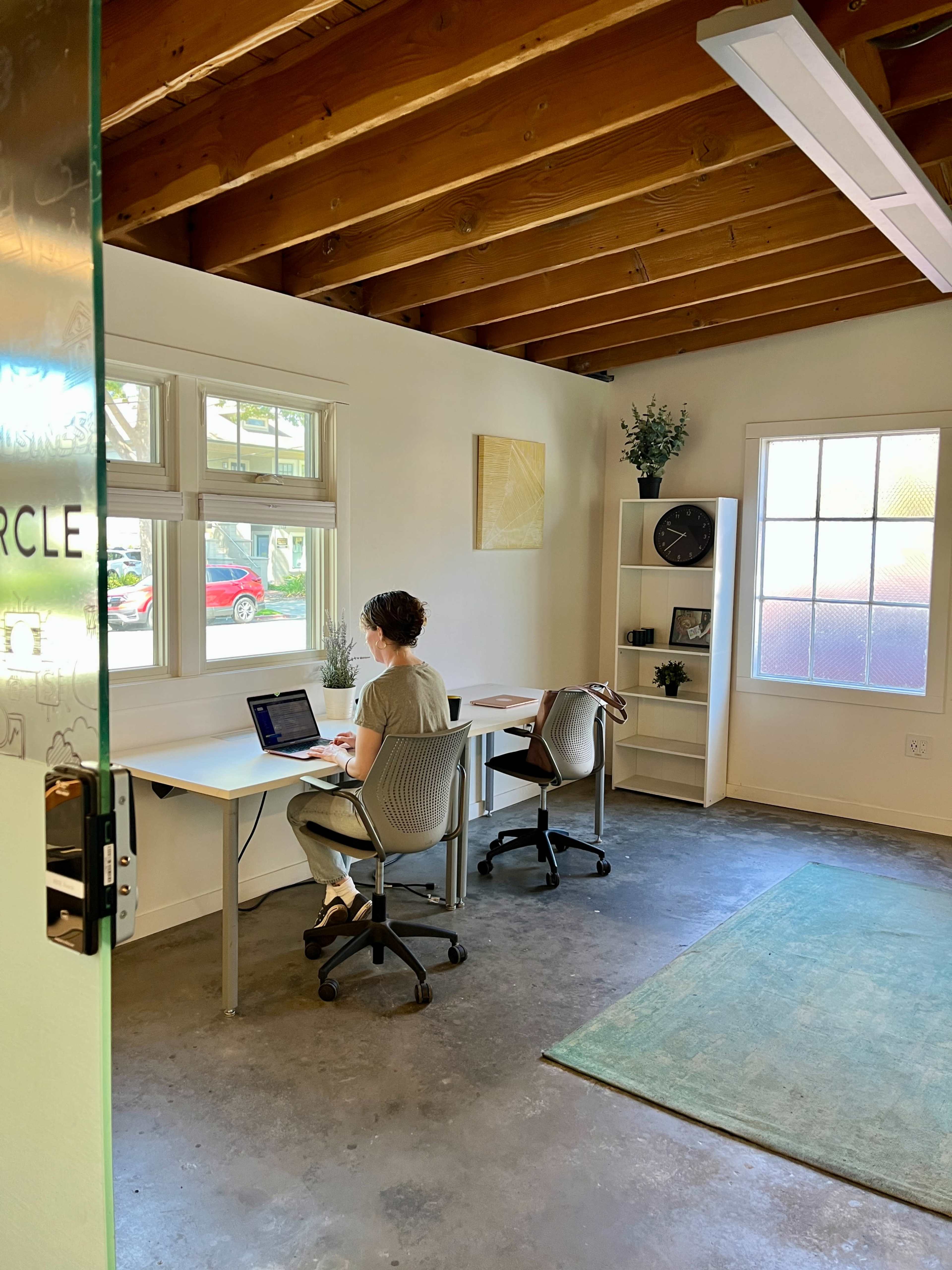 A person is sitting at a desk working on a laptop in a bright office with large windows and minimal furnishings.