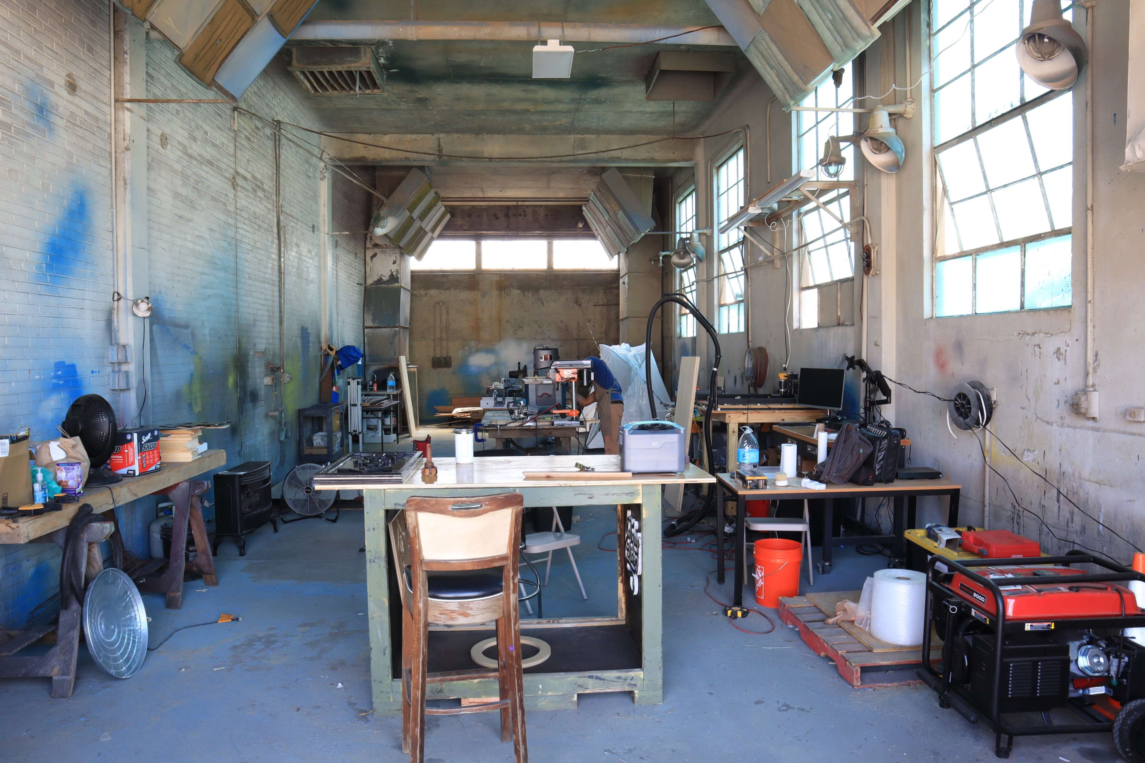 The image shows a spacious workshop with wooden tables, tools, and equipment scattered throughout, illuminated by large windows.