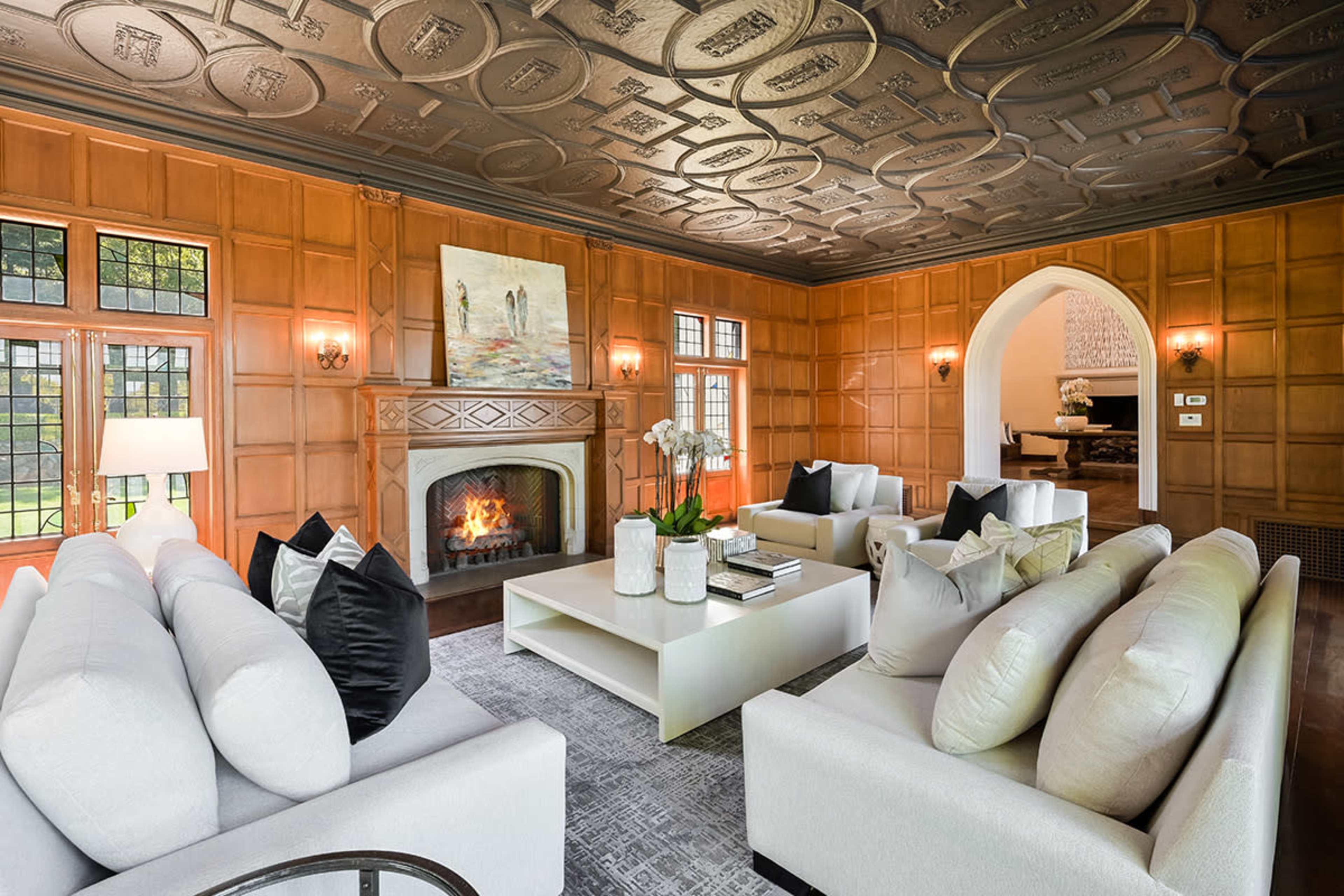 The image shows a spacious living room with wood-paneled walls, a decorative ceiling, a central fireplace, and modern white seating arranged around a coffee table.
