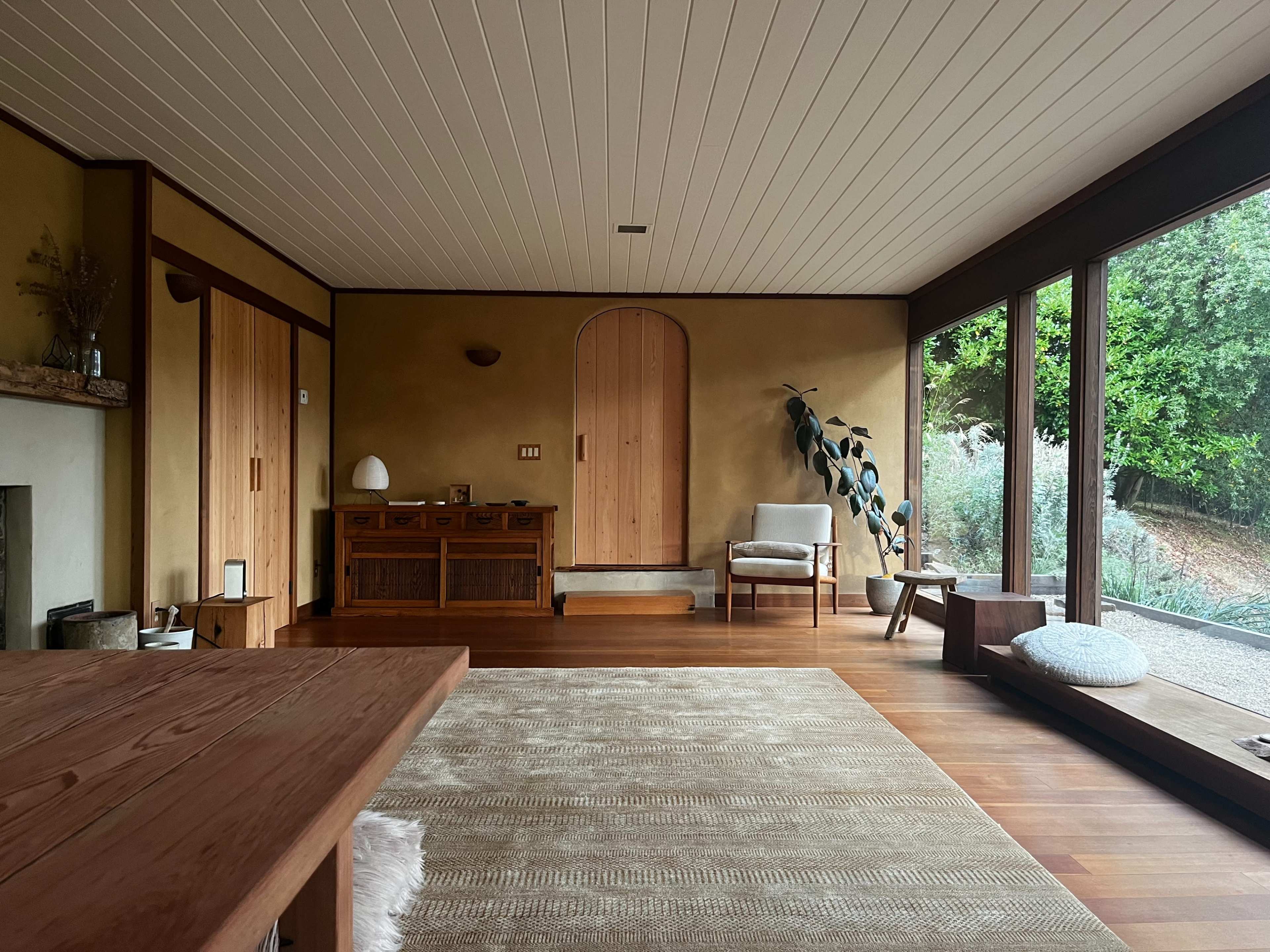 A minimalist living space with wooden furniture, a circular door, and large windows overlooking a garden.