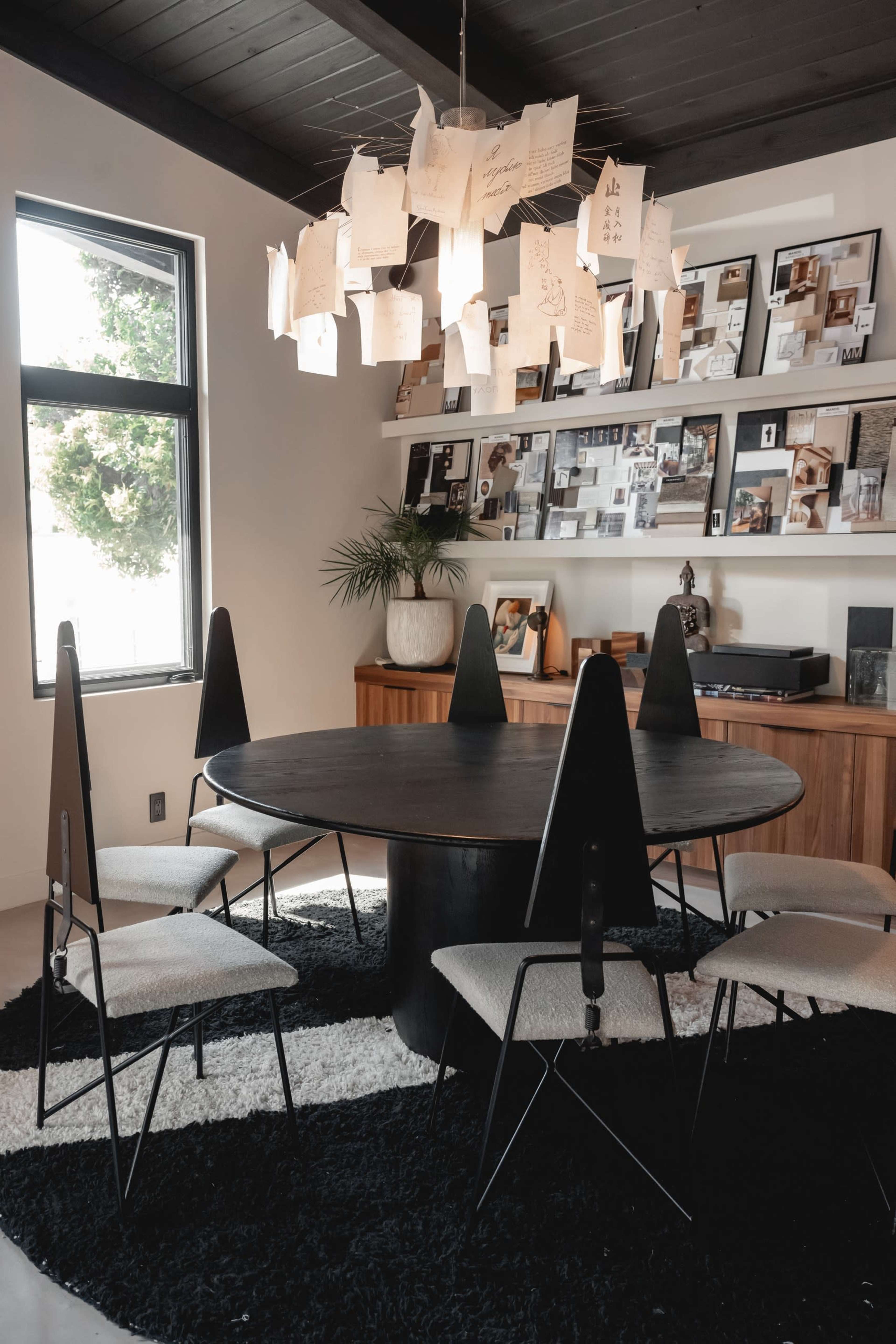 A round black table surrounded by six chairs is set in a modern room with large windows and a wall adorned with framed photographs and hanging notes.