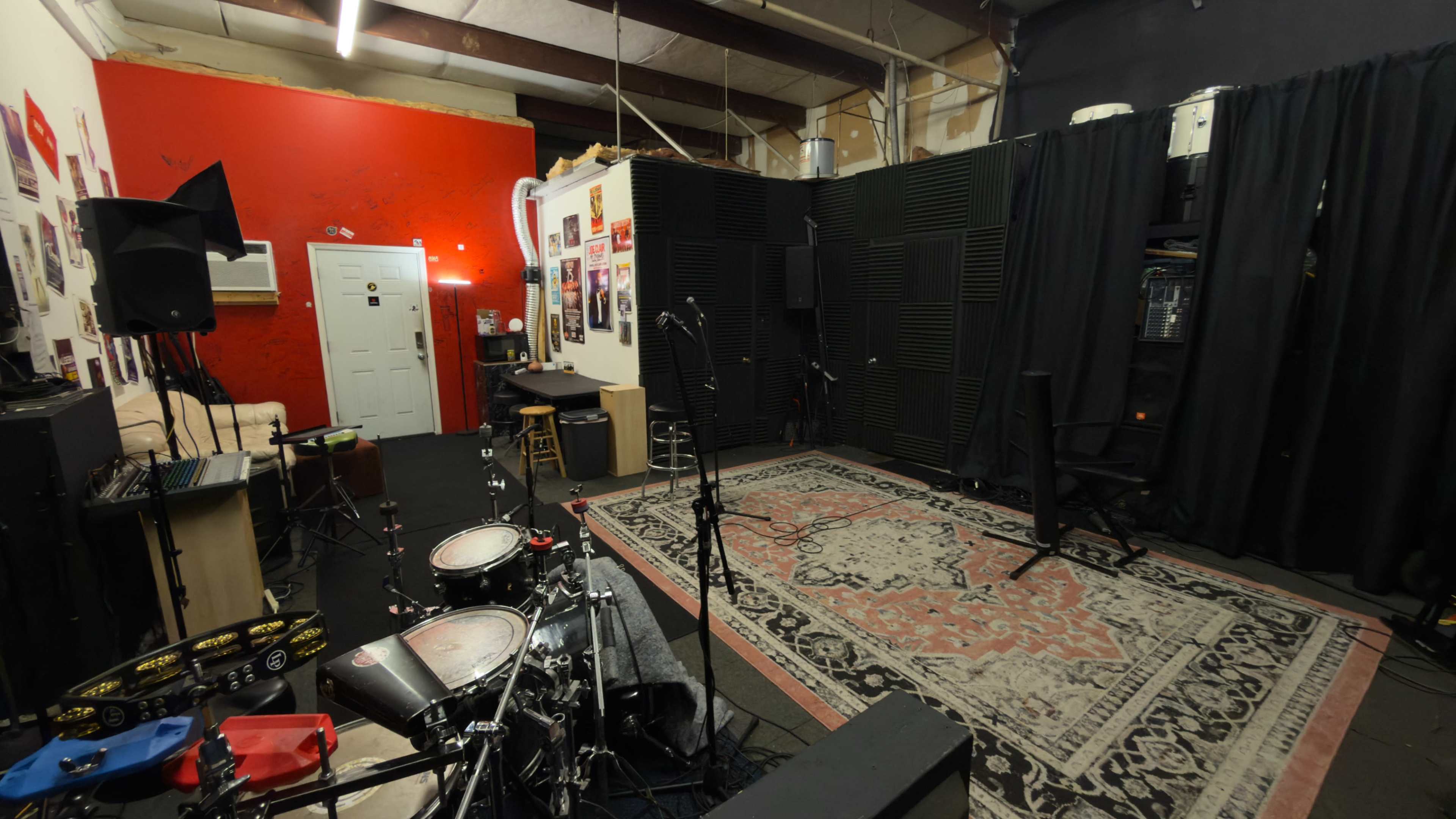 The image shows a rehearsal space with a drum set, carpet, soundproof walls, and various music equipment arranged in the room.