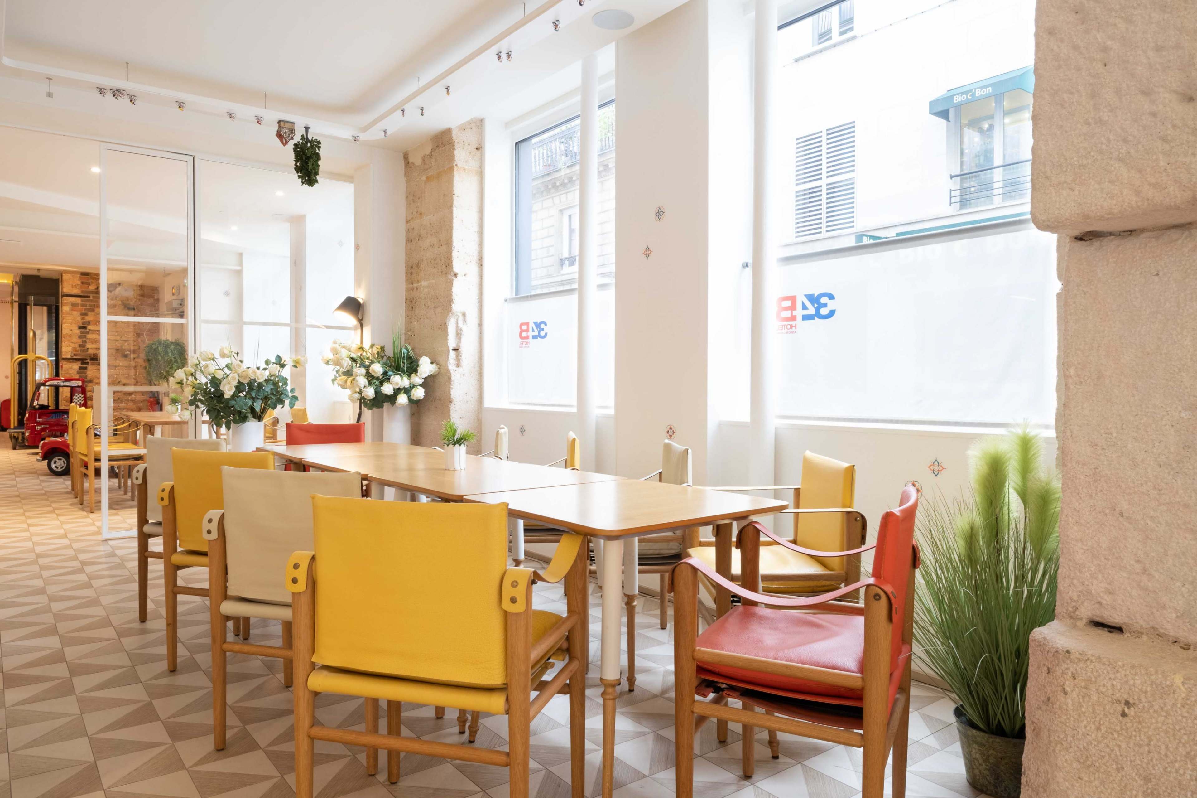 A bright dining area features a long wooden table surrounded by colorful chairs and large windows letting in natural light.