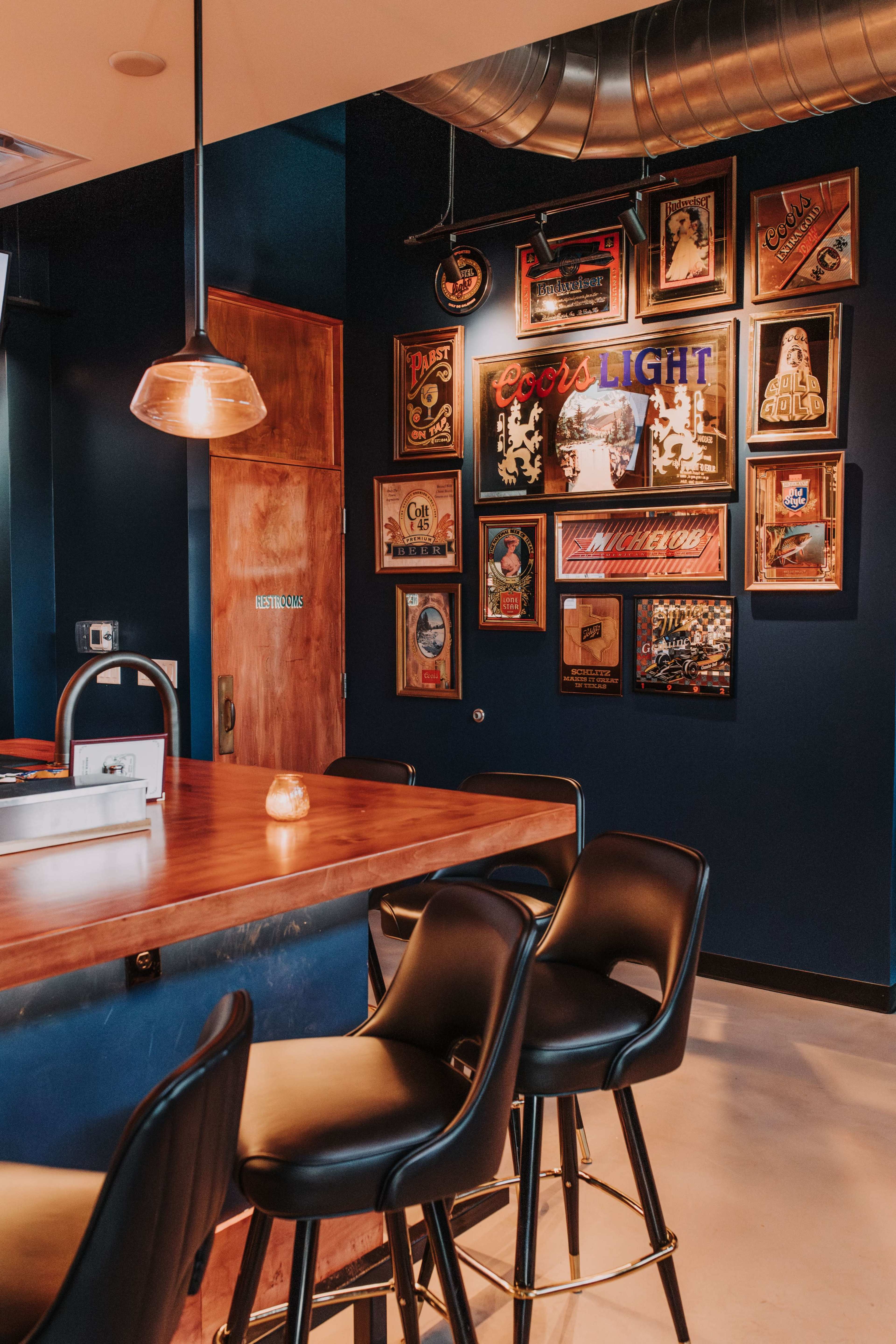 The image shows a bar area with a wooden counter, black stools, and a wall adorned with framed vintage advertisements.