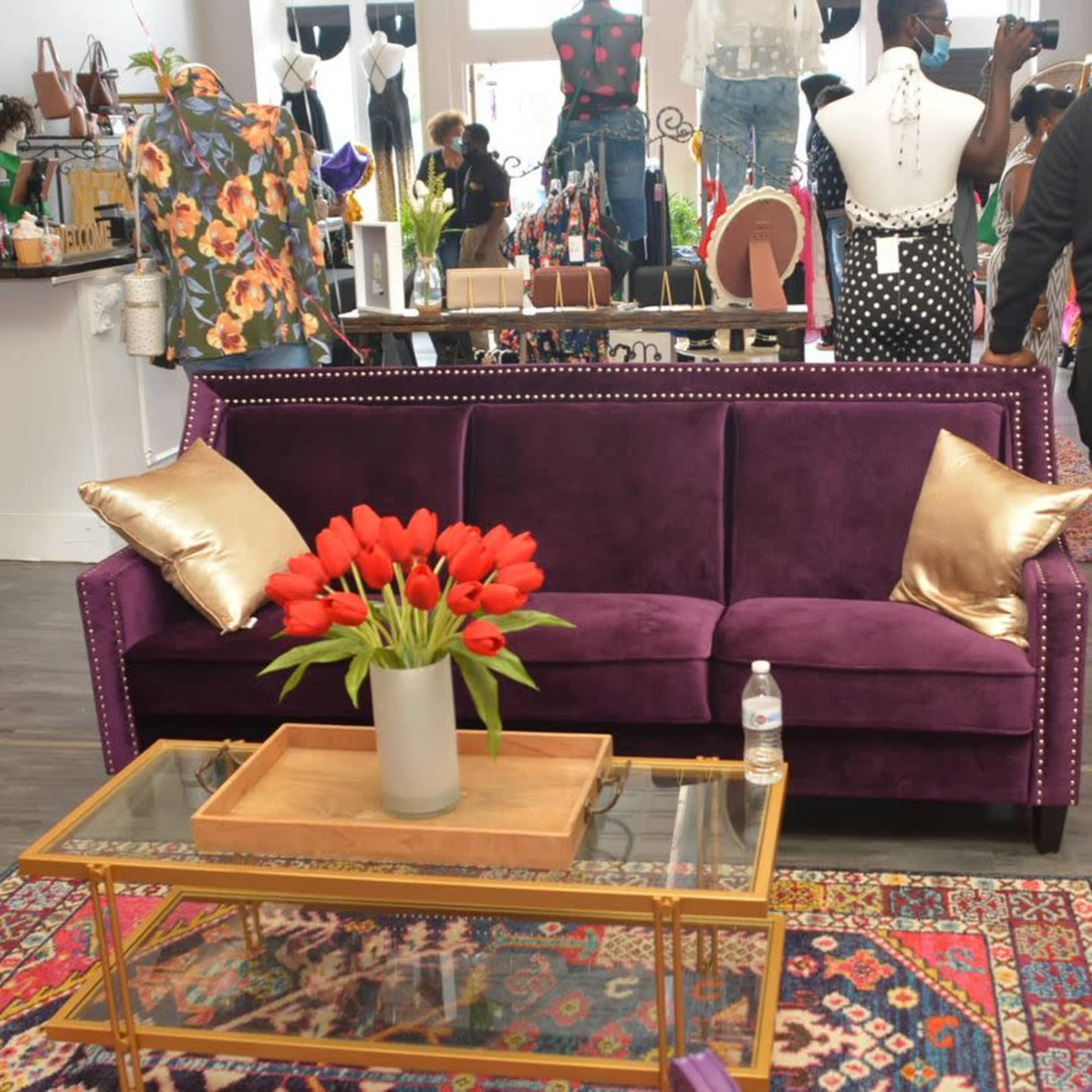 A purple couch with decorative pillows is positioned in front of a glass coffee table, adorned with a vase of red tulips, within a boutique displaying various clothing items in the background.