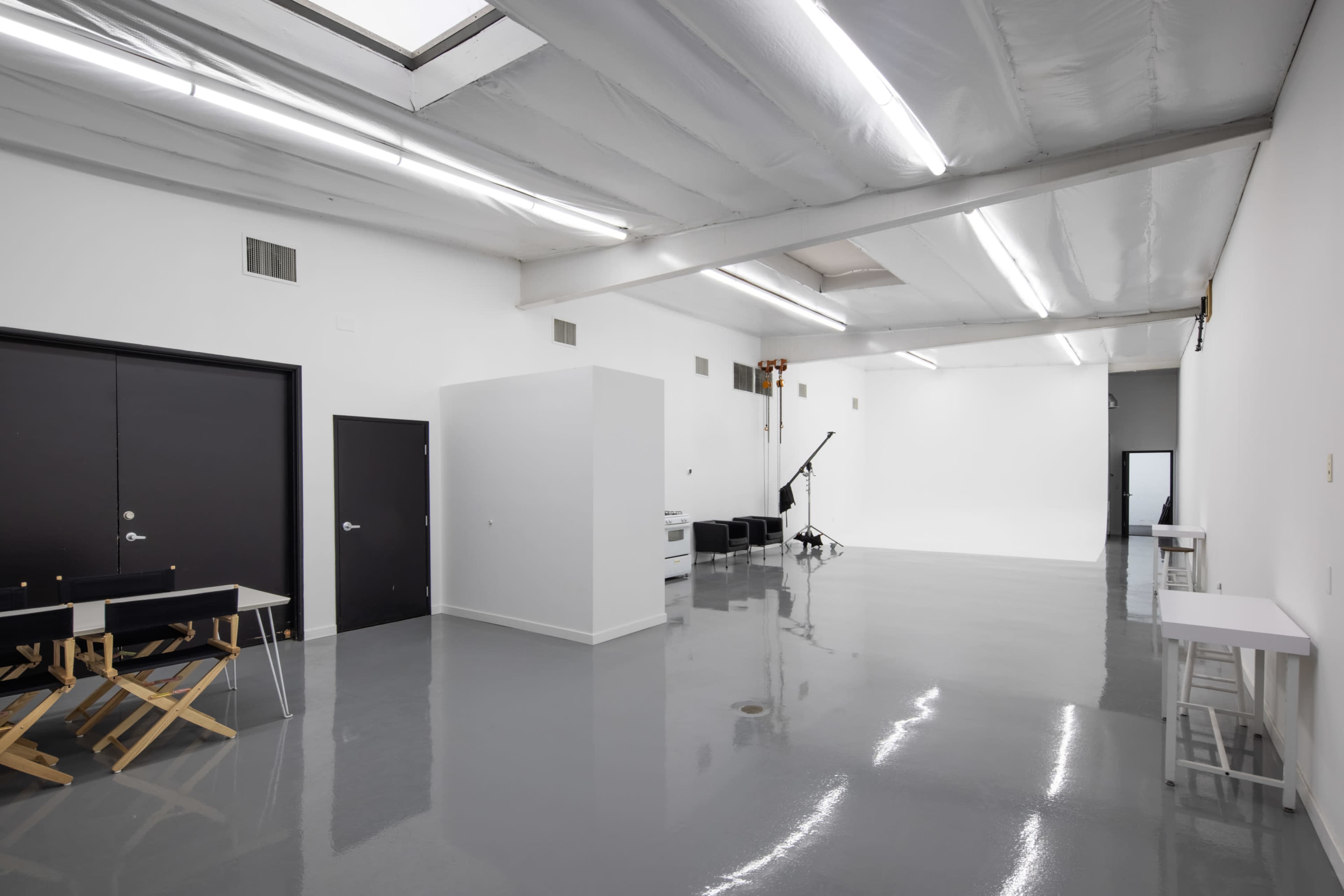 A spacious, brightly lit studio with polished concrete flooring, featuring black doors, a white partition, and a backdrop area.