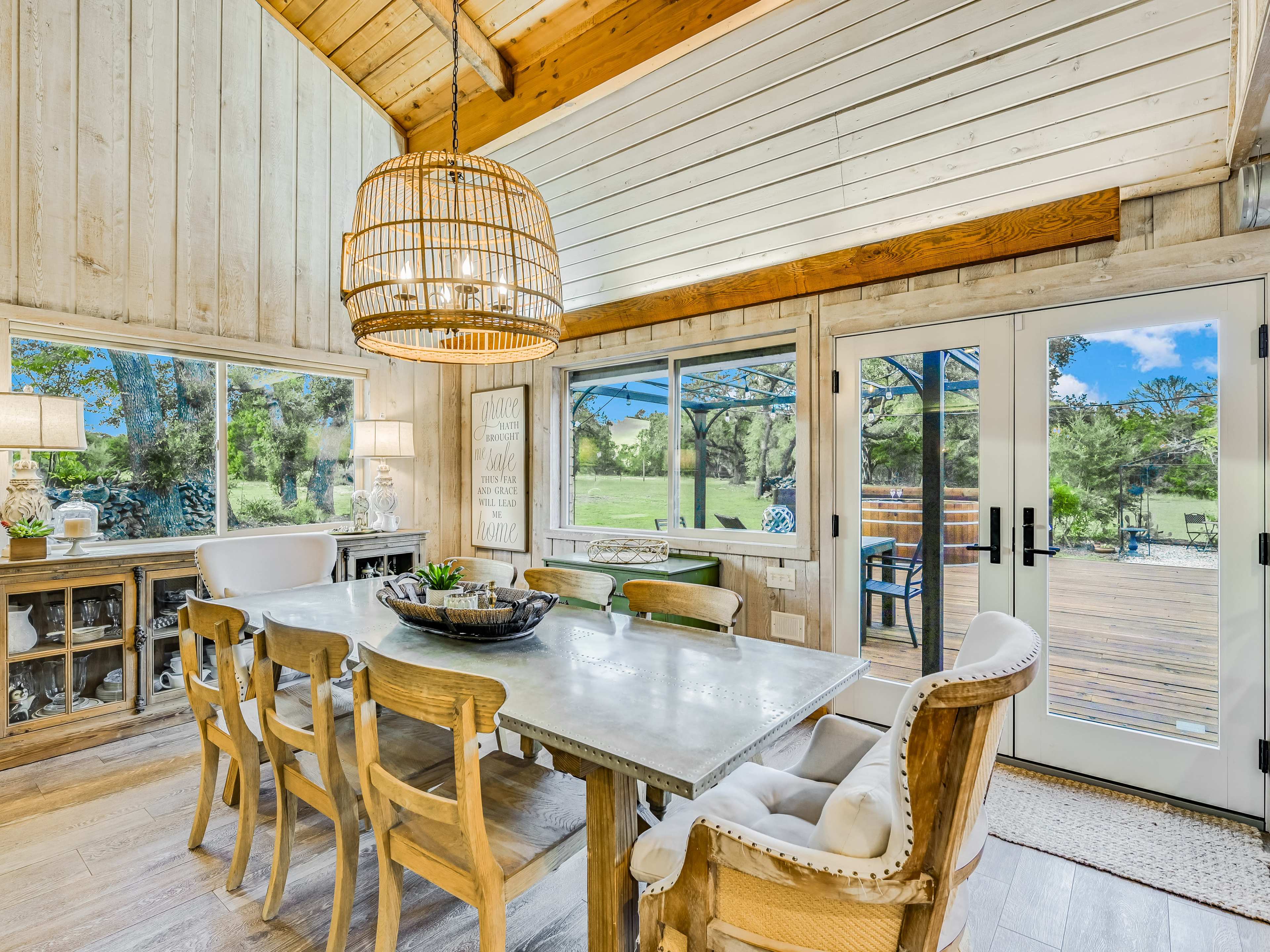 A dining area features a large table surrounded by chairs, with a woven pendant light above and doors leading to an outdoor deck.