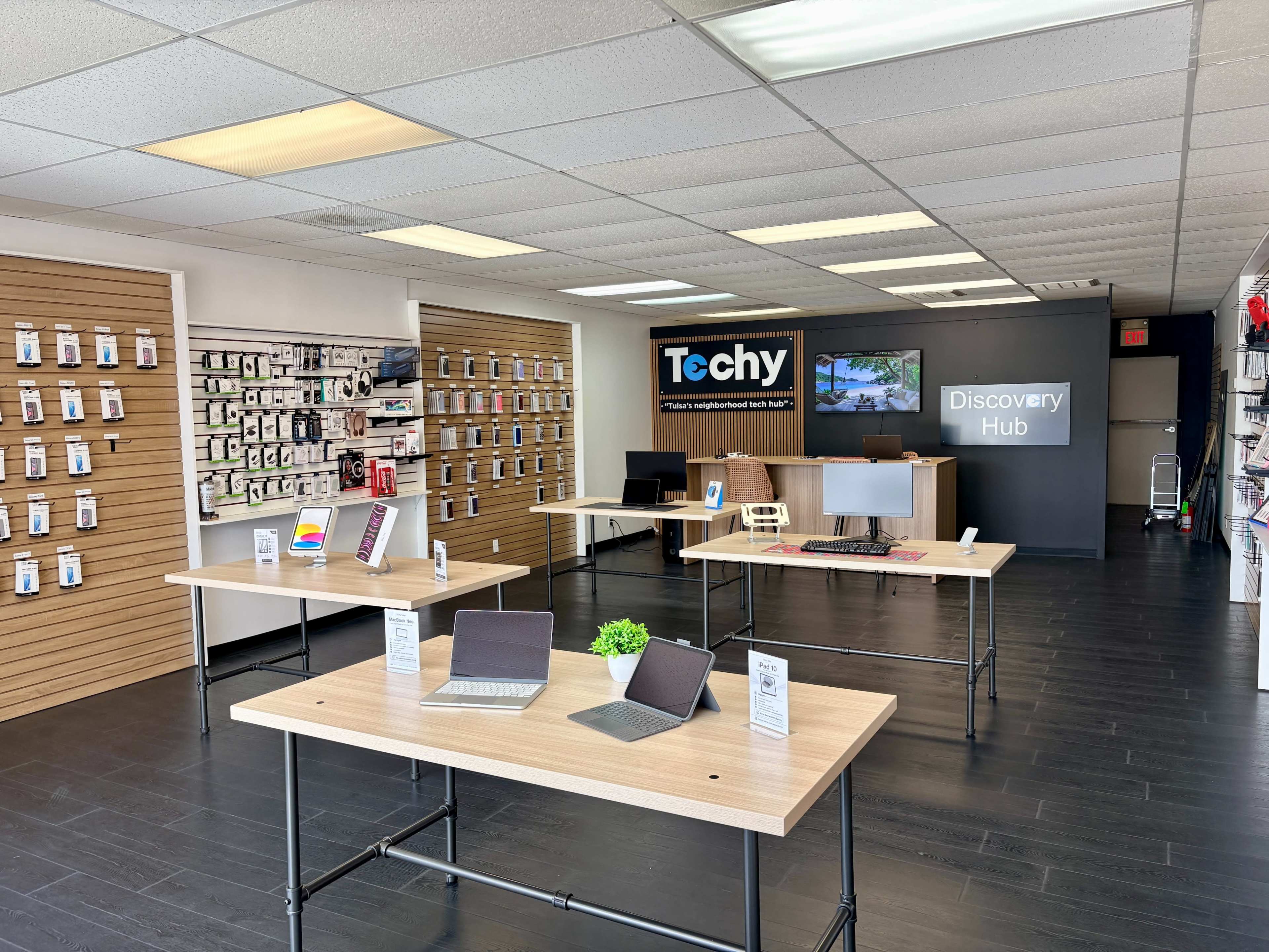The image shows an electronics retail store with displays of gadgets and devices on the walls and several wooden tables arranged throughout the space.