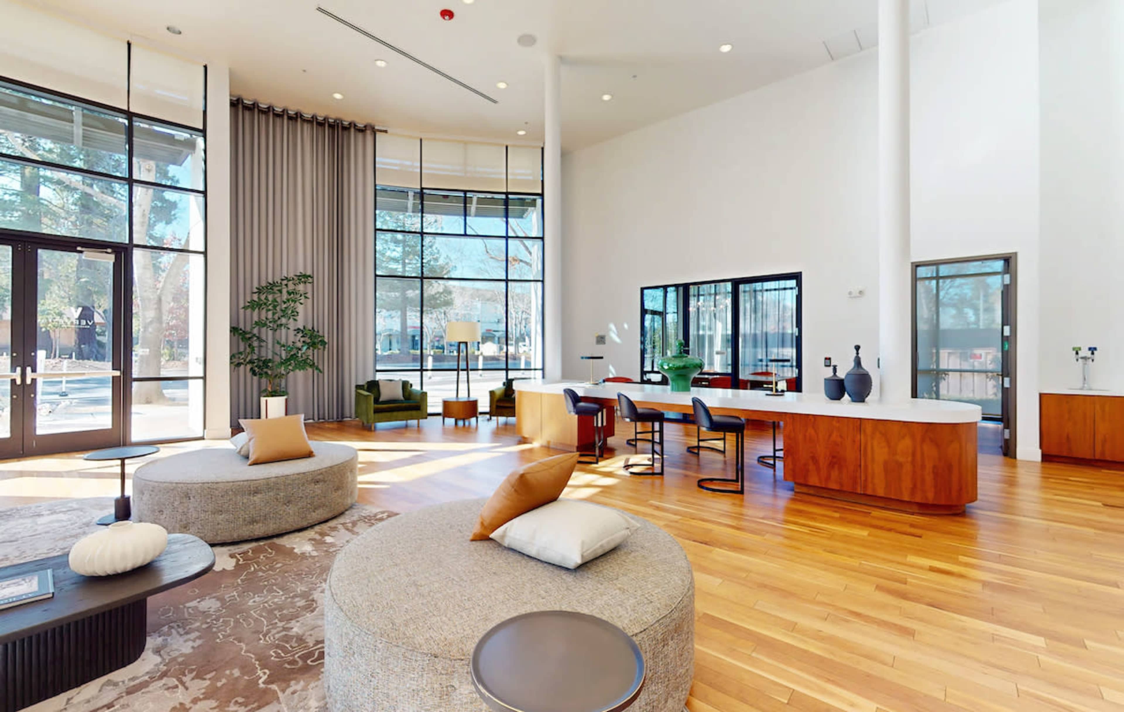 The image shows a modern lounge area with large windows, round seating cushions, a light wood floor, and a stylish bar counter.