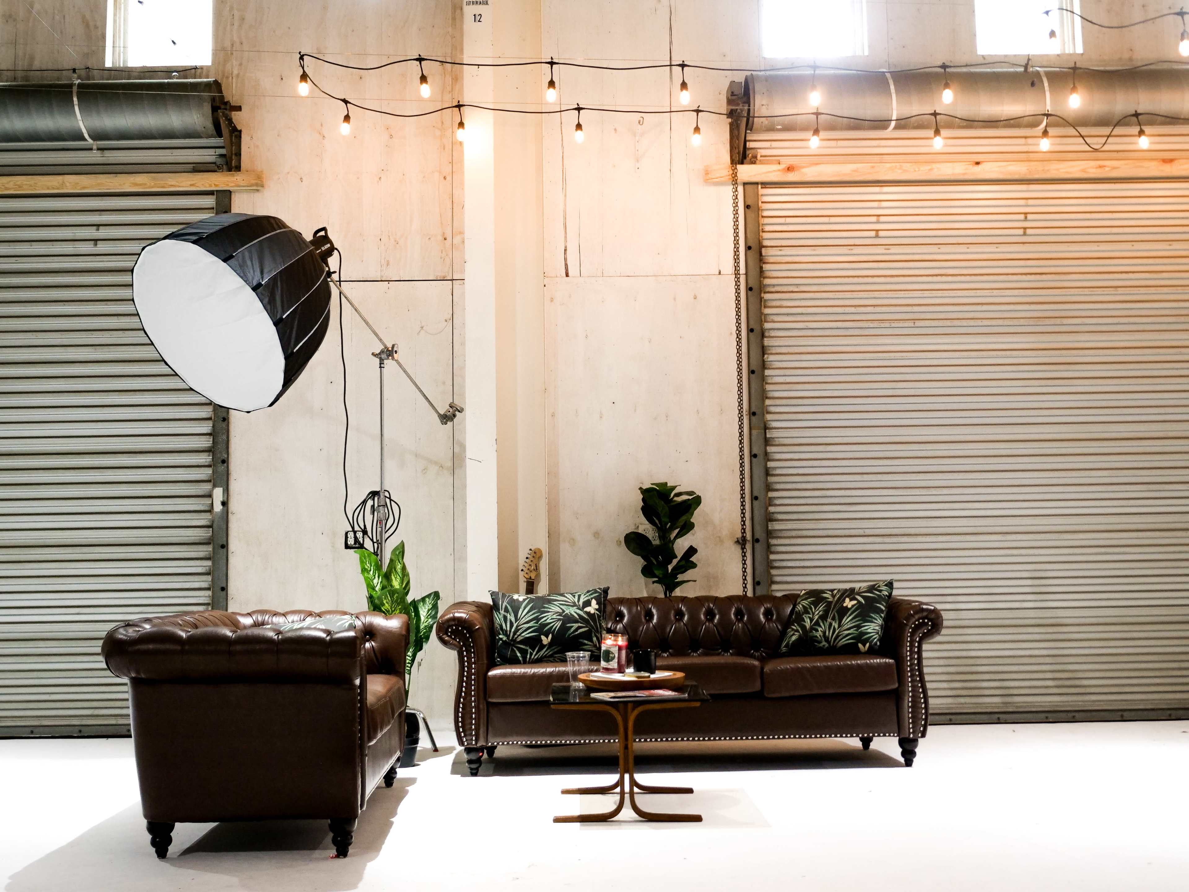 A stylish indoor space featuring two brown leather sofas, a round wooden table with drinks, and decorative plants, illuminated by hanging light bulbs.
