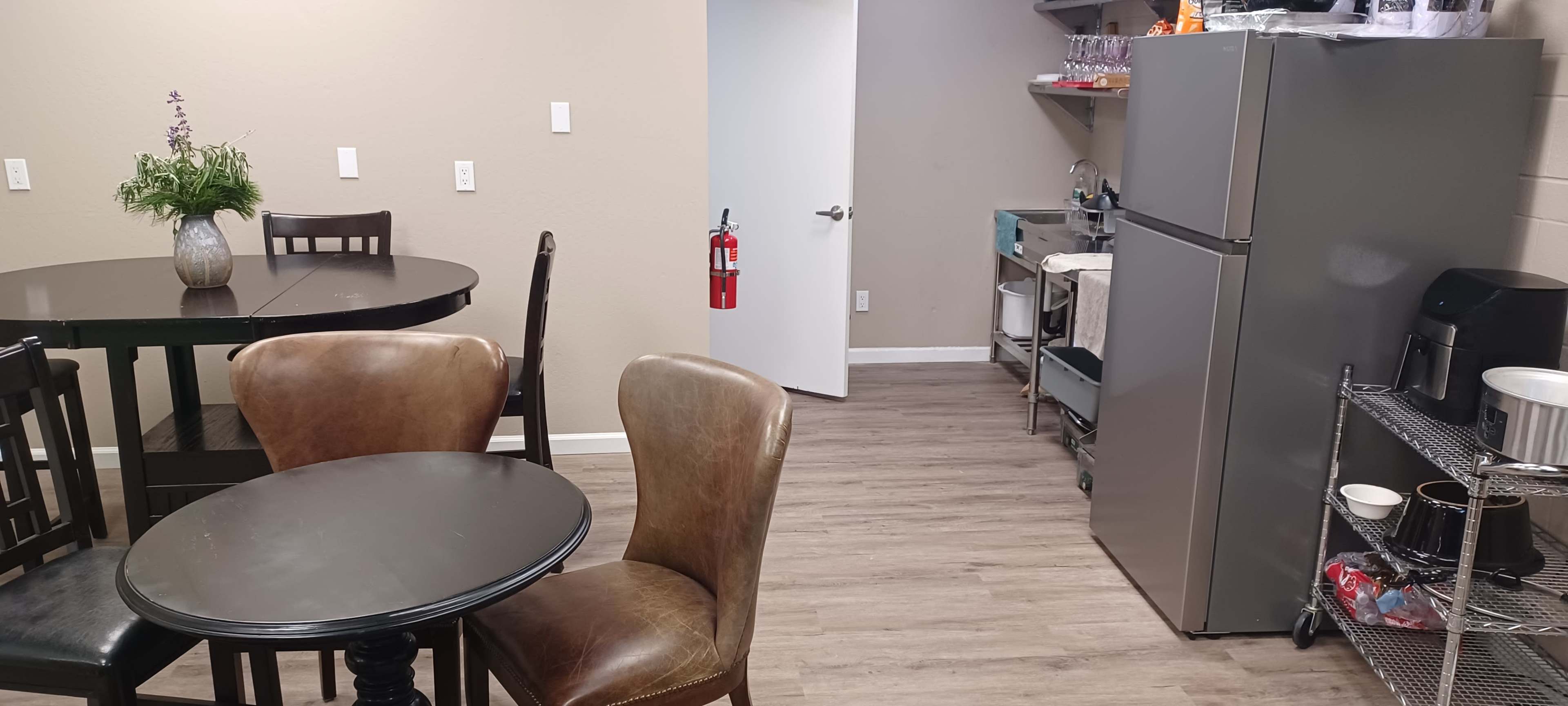 The image shows a small break room featuring two round tables and chairs, a kitchen area with a refrigerator and shelving, and a door leading to another space.