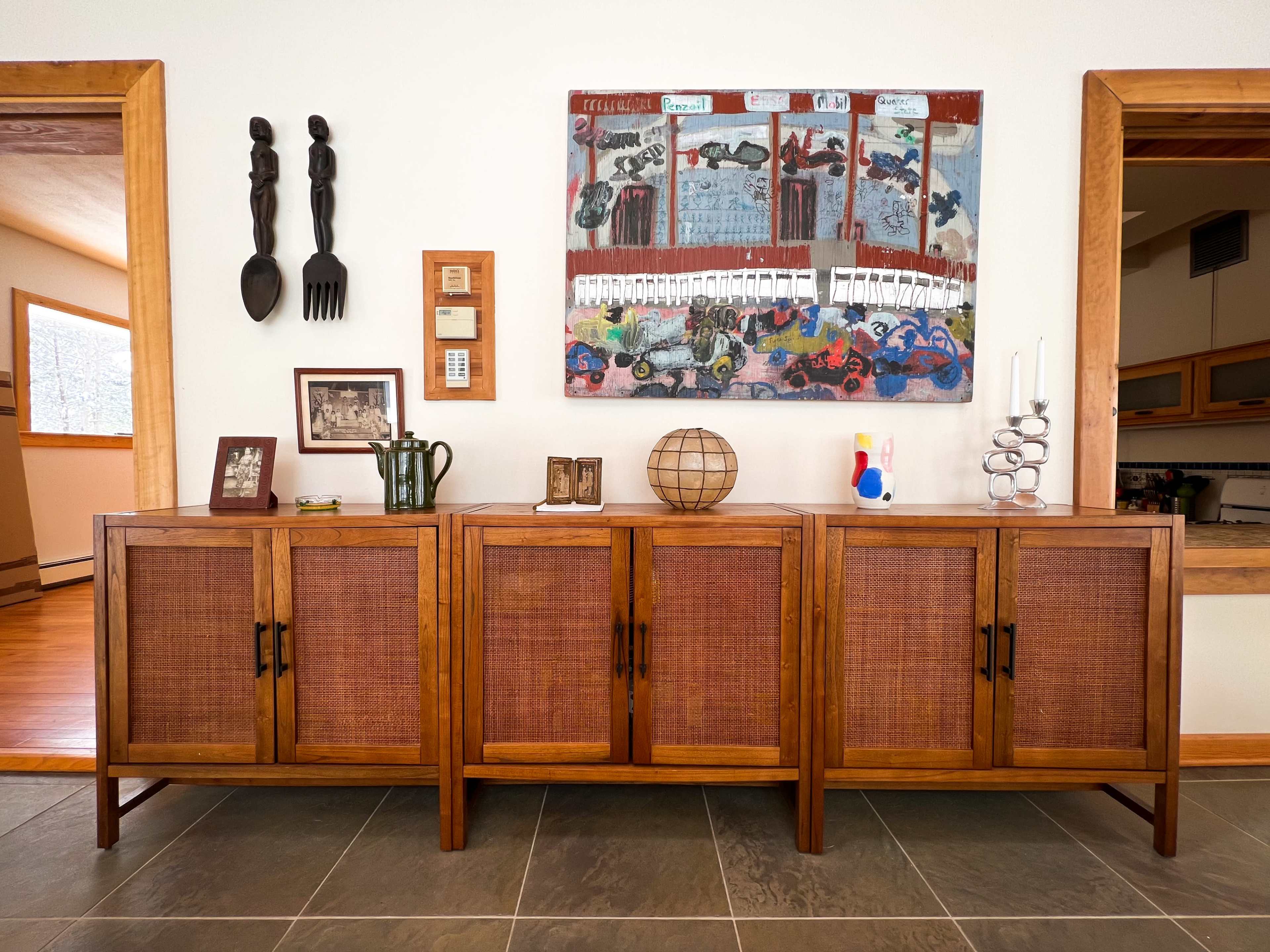 A wooden sideboard with woven cabinet doors, decorative art on the wall, and various objects arranged on top, including framed photos and a globe.
