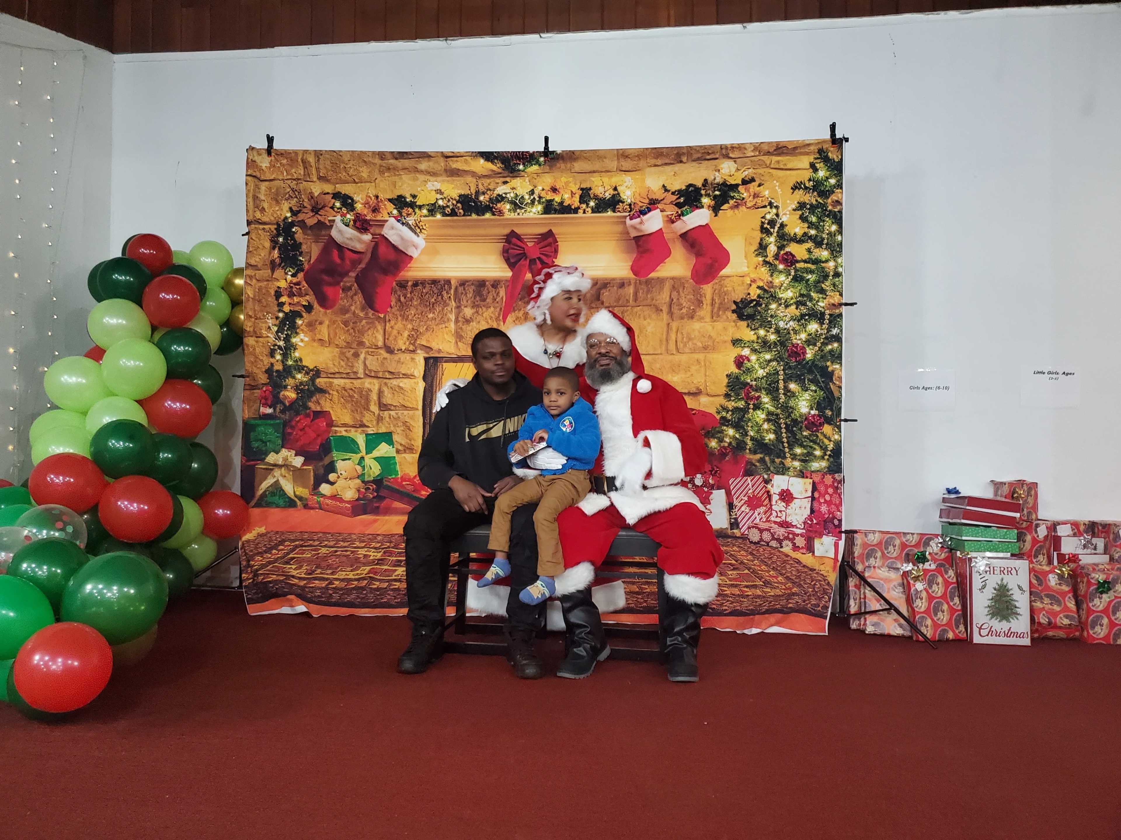 A man and a young boy sit on a chair with Santa Claus in front of a festive backdrop featuring Christmas decorations and gifts.
