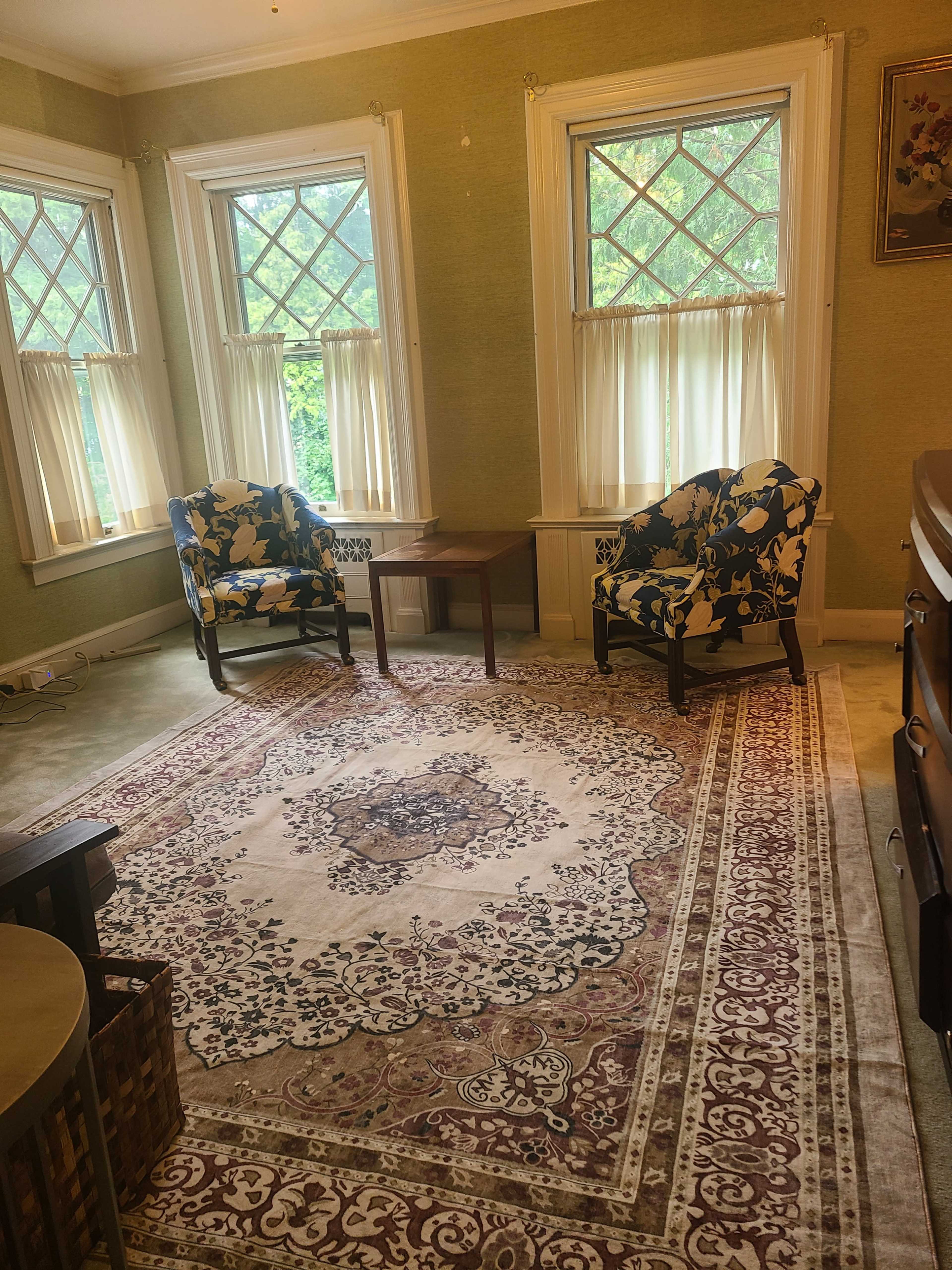 The image shows a cozy room featuring two patterned chairs, a small table between them, and large windows adorned with sheer curtains, all set over a decorative area rug.