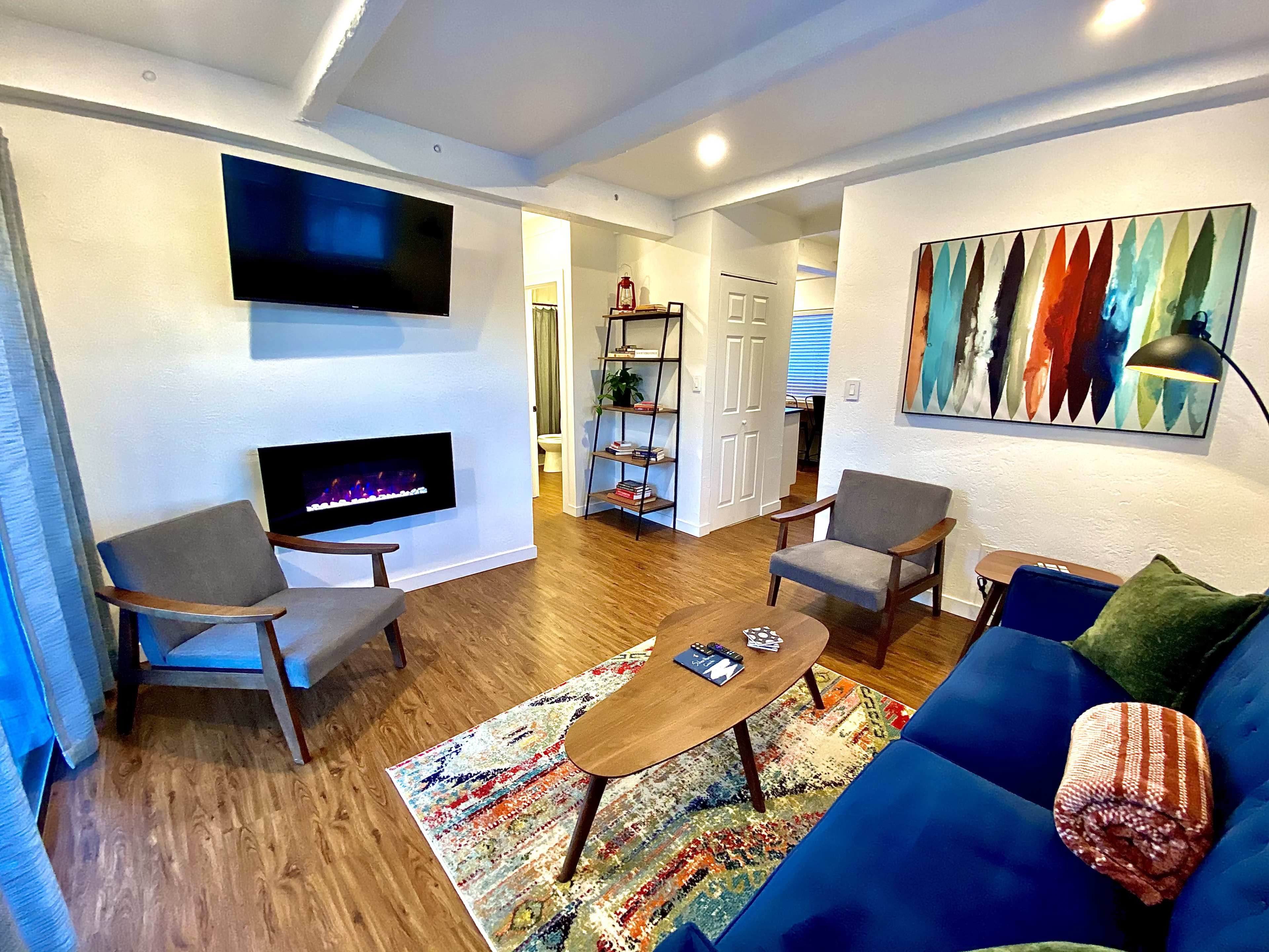 The living room features a blue couch, two gray chairs, a wooden coffee table on a patterned rug, a wall-mounted TV, and an electric fireplace.